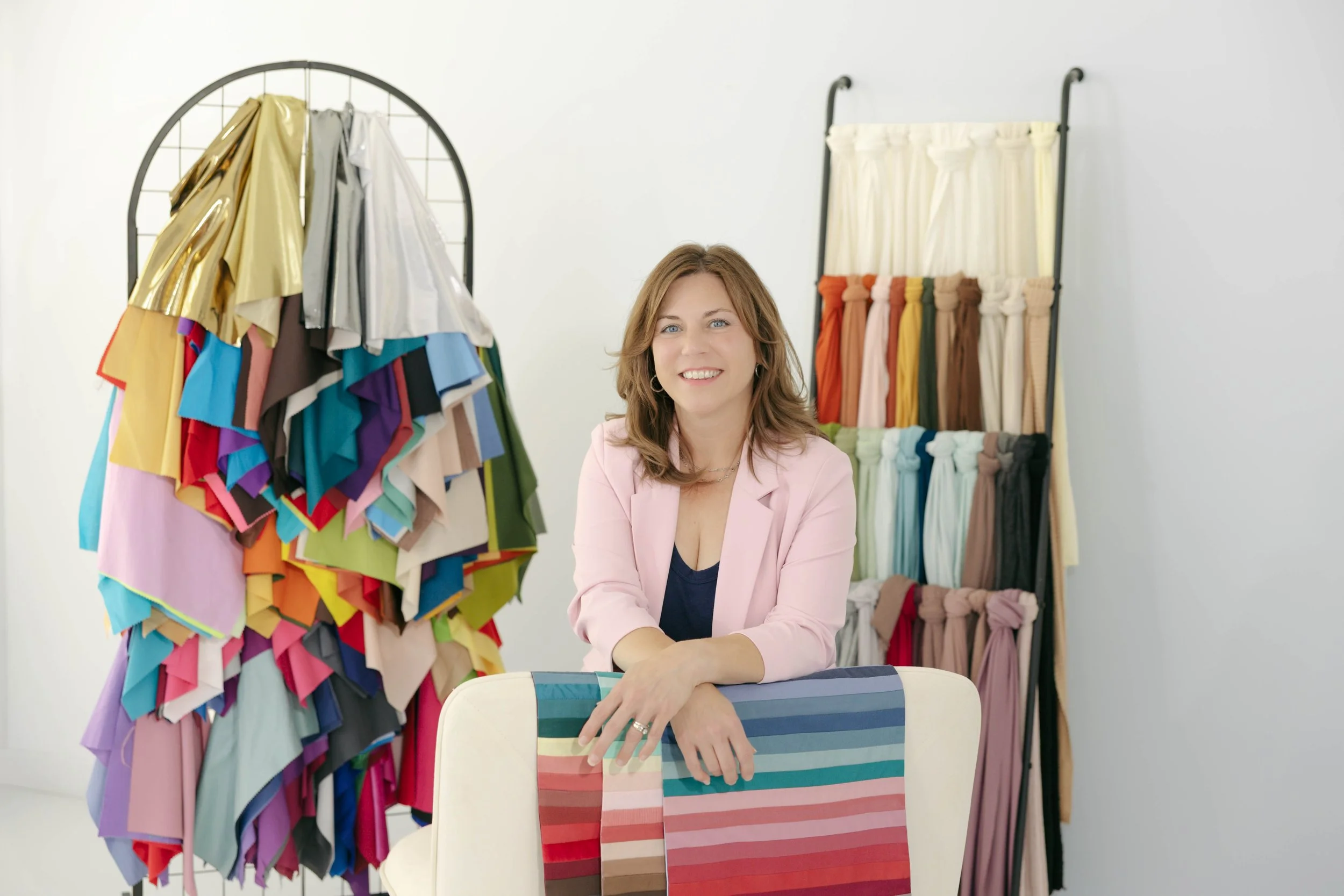 A woman sits at a table in front of racks filled with fabric swatches and clothing material samples in a bright, modern fabric store or showroom.