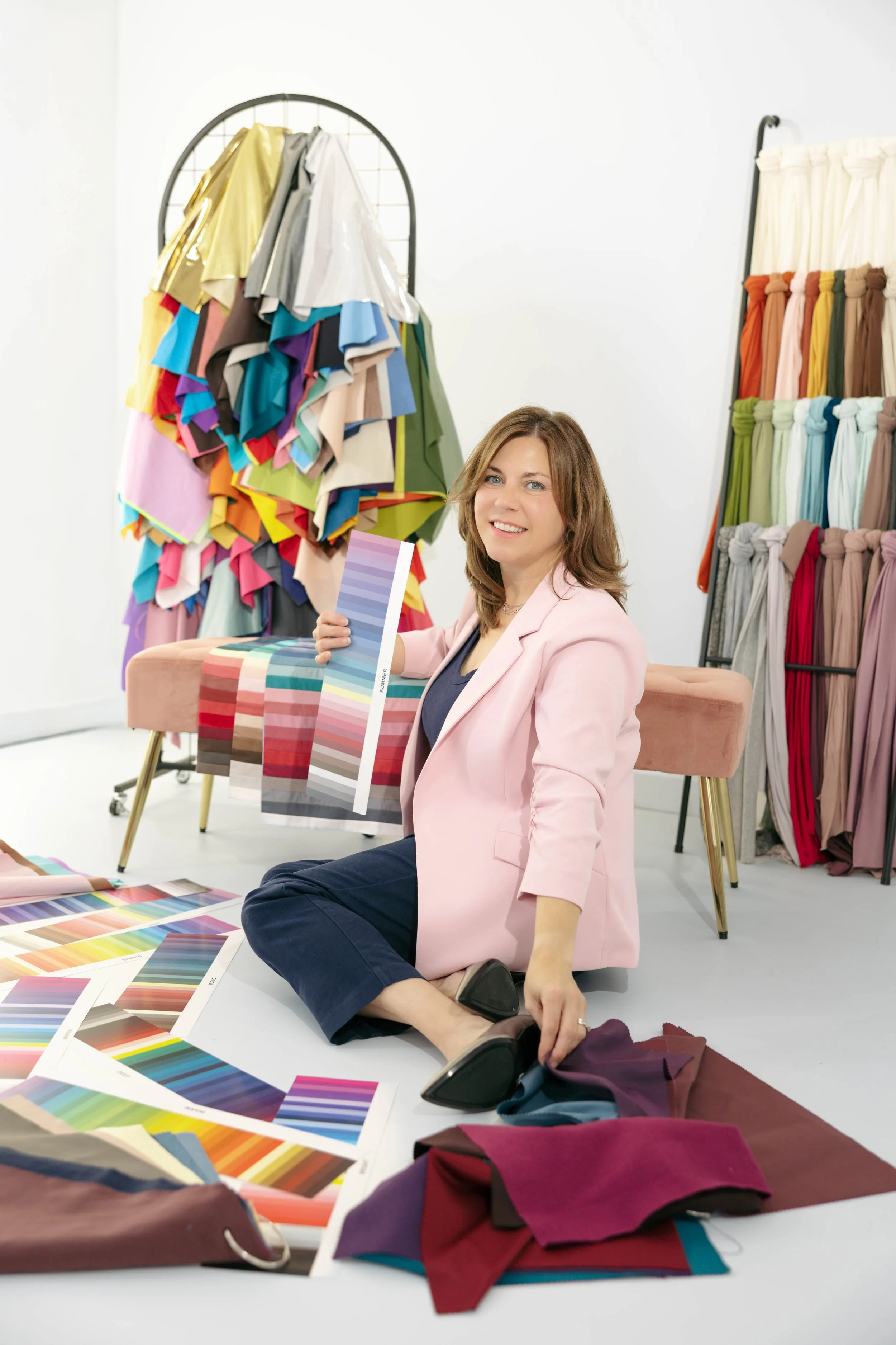 A woman sitting on the floor surrounded by fabric swatches and color samples in a bright room with color swatches and fabric samples in the background.