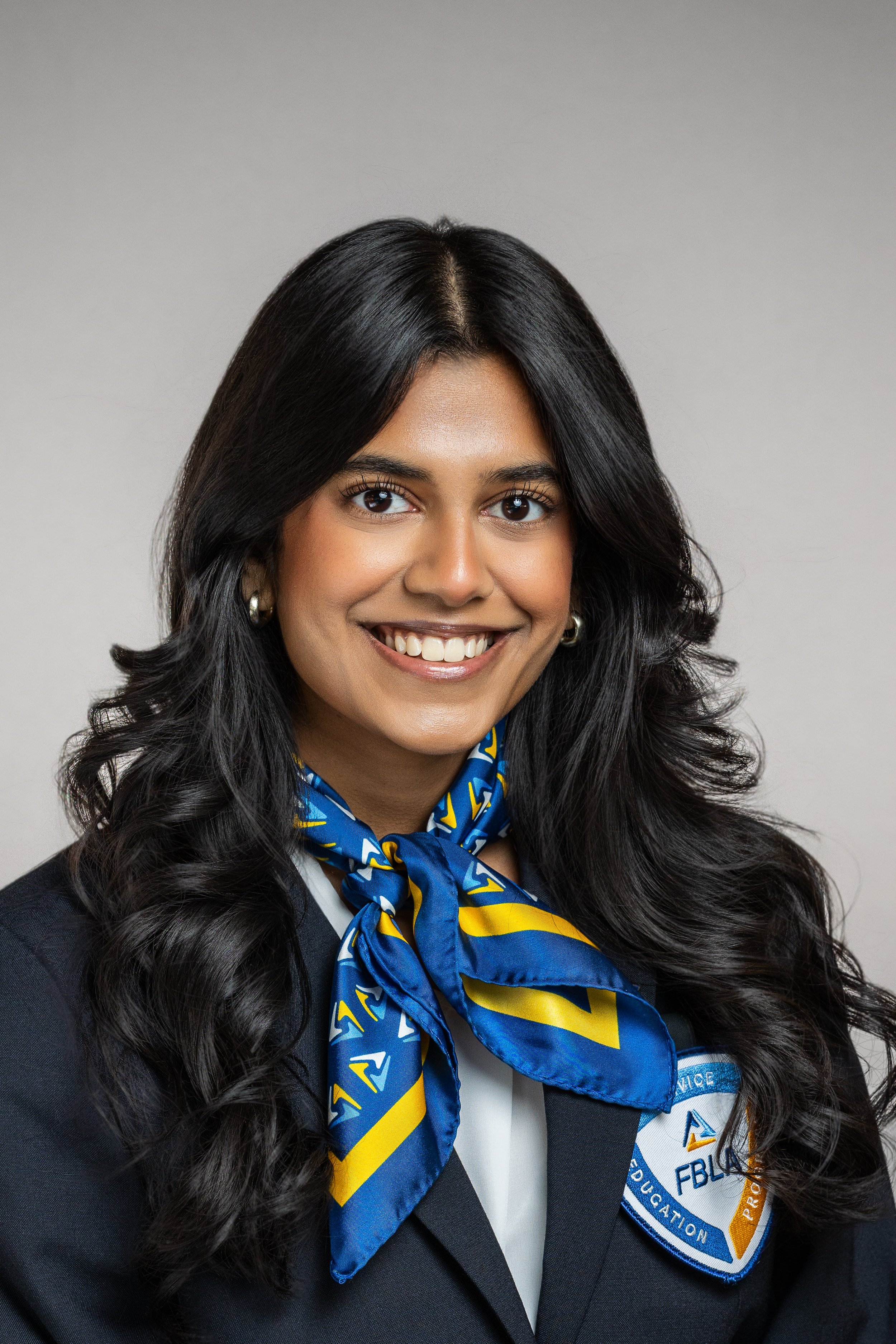 Woman with long dark hair wearing a black blazer, white shirt, blue and yellow patterned scarf, and a badge with the logo of the Future Business Leaders of America (FBLA).