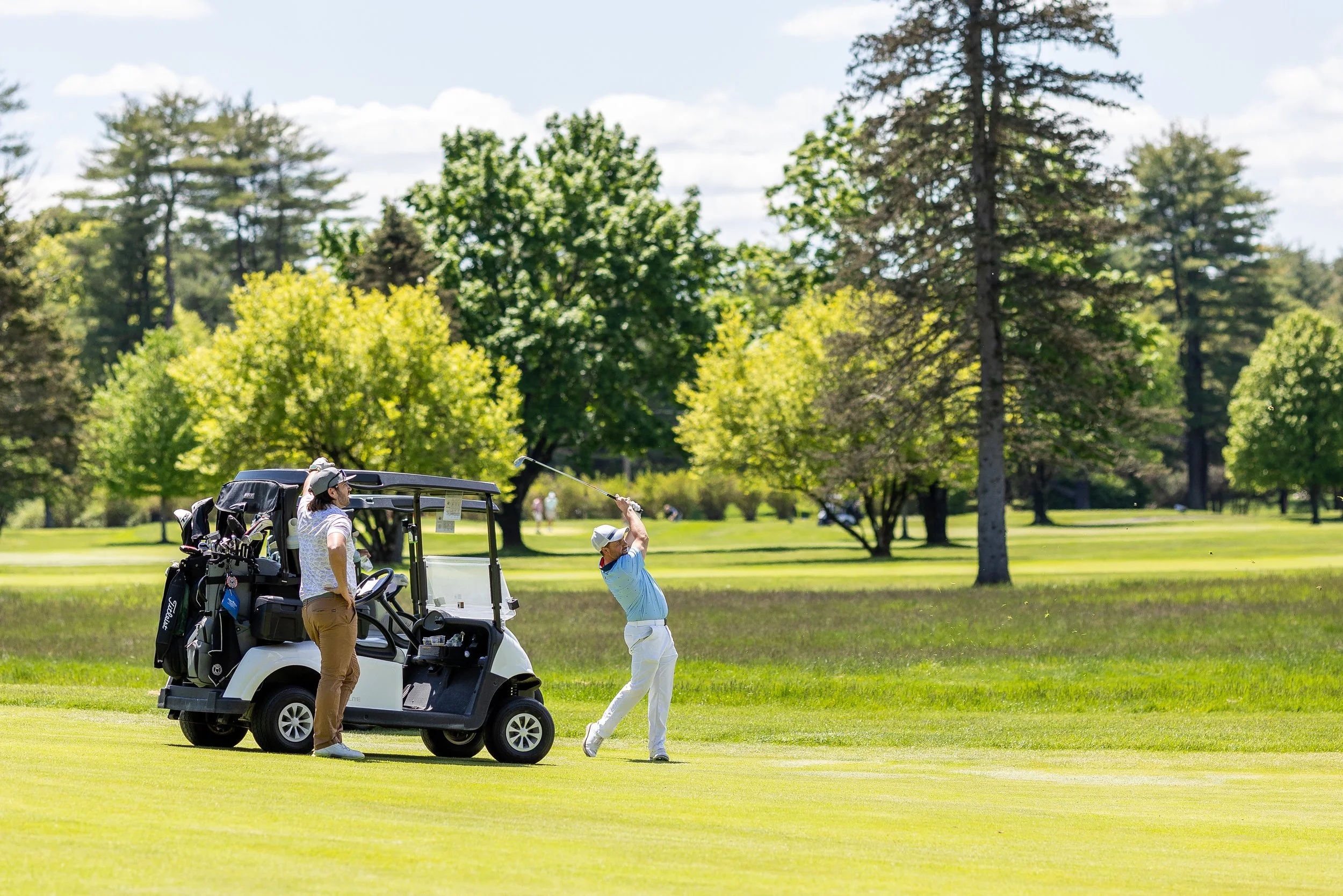 A person in a blue shirt and white pants swinging a golf club on a golf course with a golf cart nearby, and another person standing beside it. The background features lush green trees and a sunny sky.