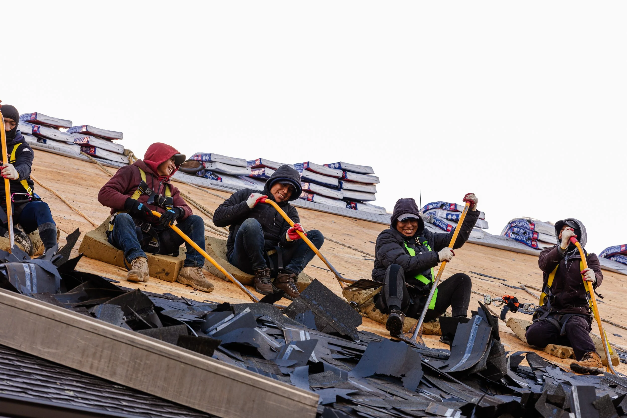 Roofing workers installing shingles on a roof, wearing safety gear and using tools, with bundles of shingles in the background.