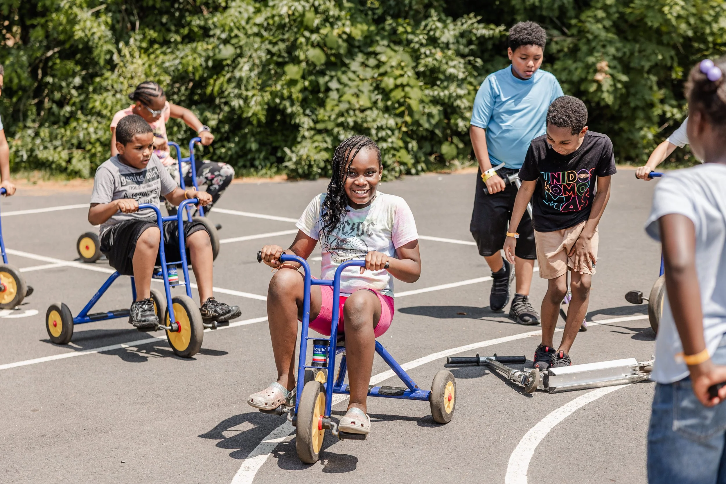Children playing on outdoor tricycles and scooters on a paved court with green bushes in the background.