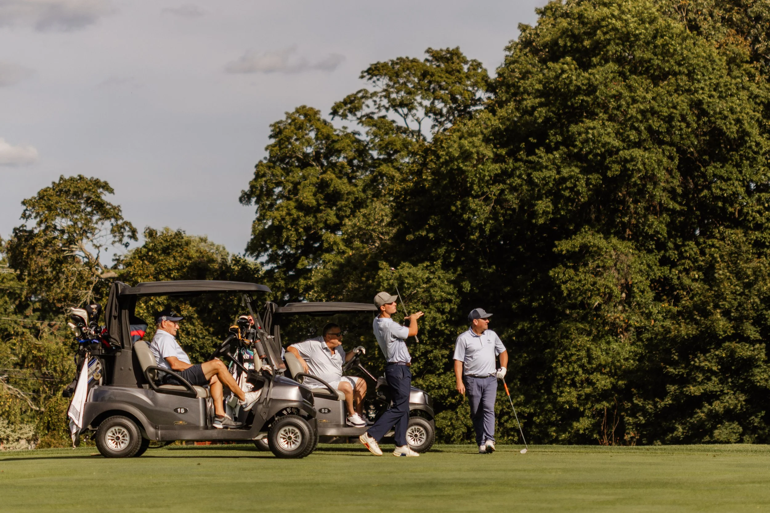Group of people playing golf on a golf course with two golf carts and lush trees in the background.