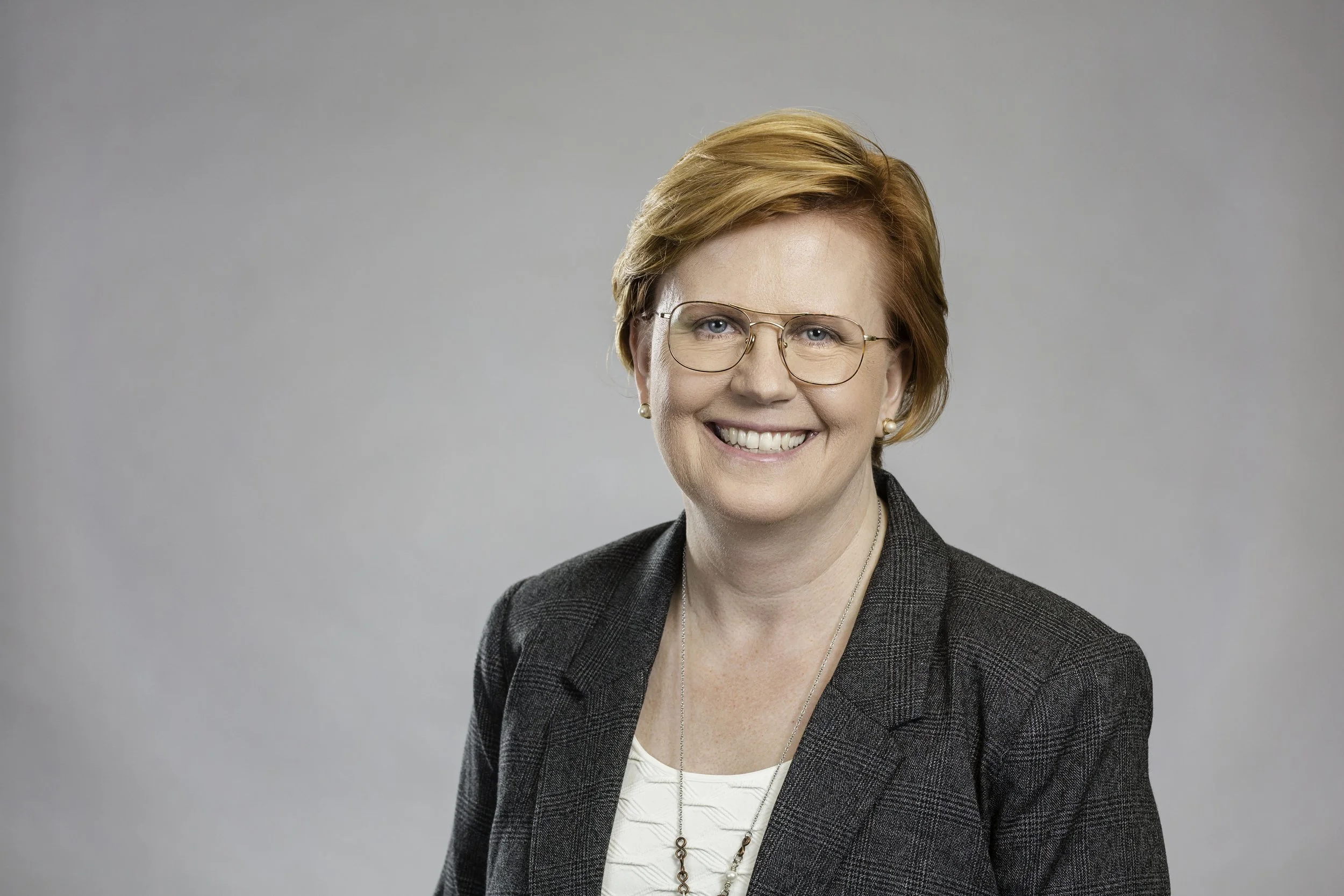 Professional woman with short red hair and glasses smiling, dressed in a dark blazer and light blouse, against a neutral background.