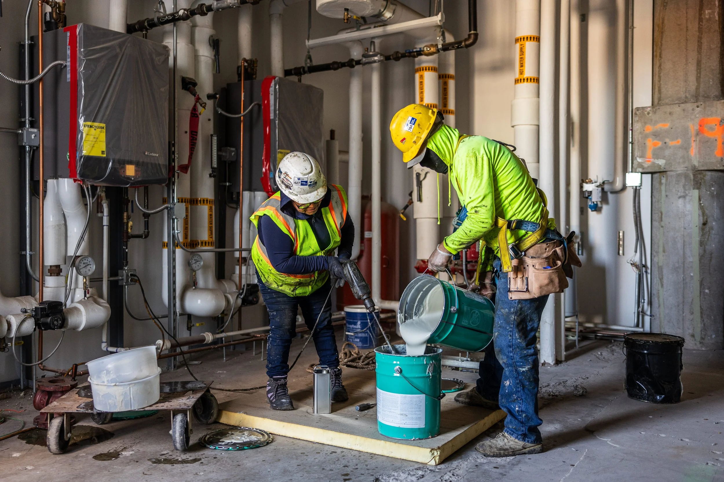 Two construction workers in safety gear working with a drill and pouring sealant or coating into a container on a construction site.