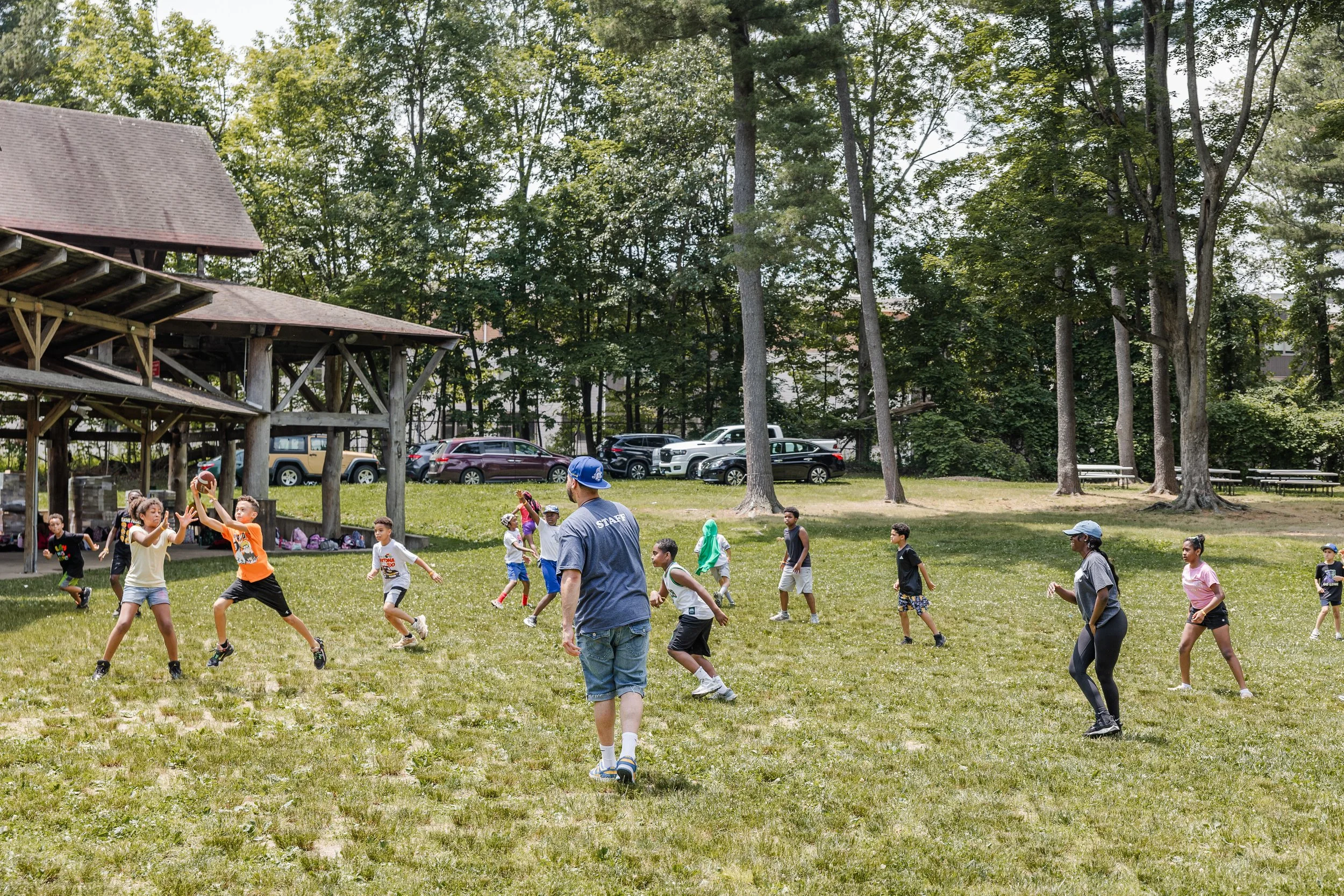 Children playing catch with a football on a grassy field with adults supervising, trees, parked cars, and a wooden pavilion in the background.