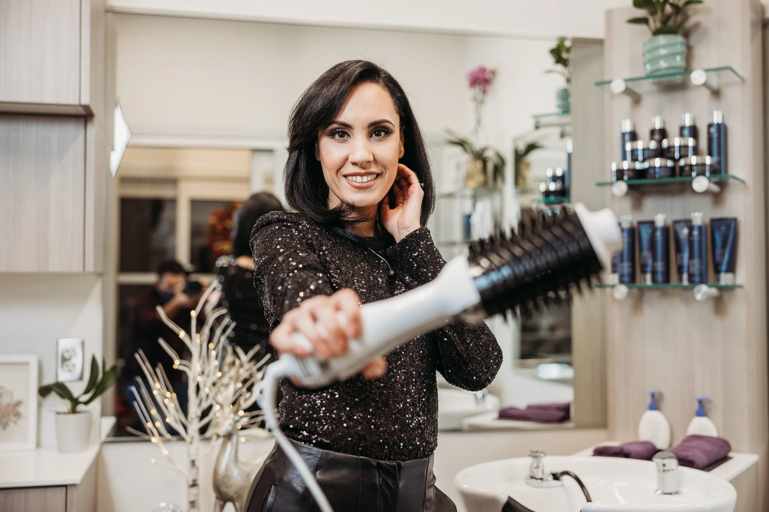 A woman smiling and holding a round hairbrush in a salon.