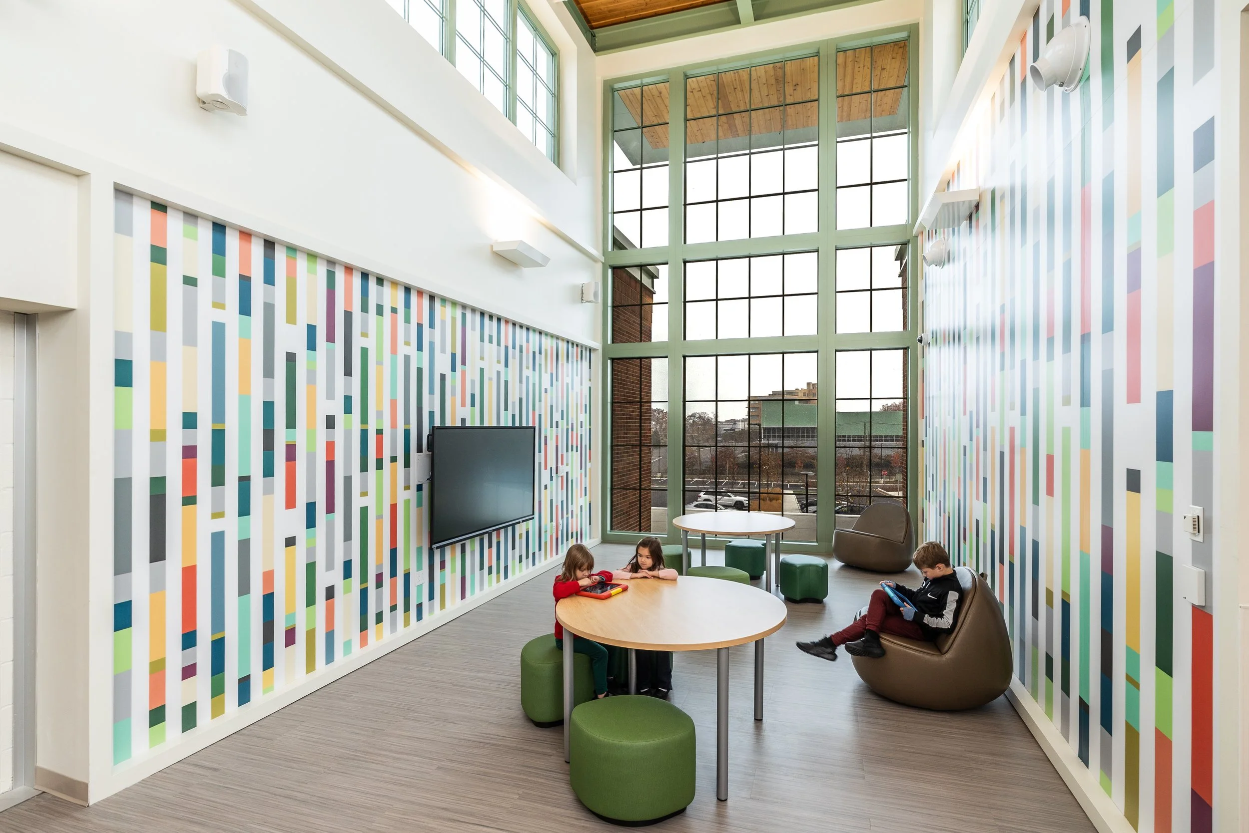A modern interior space with colorful striped wall design, large floor-to-ceiling windows, and children sitting on green and brown furniture using tablets and books.