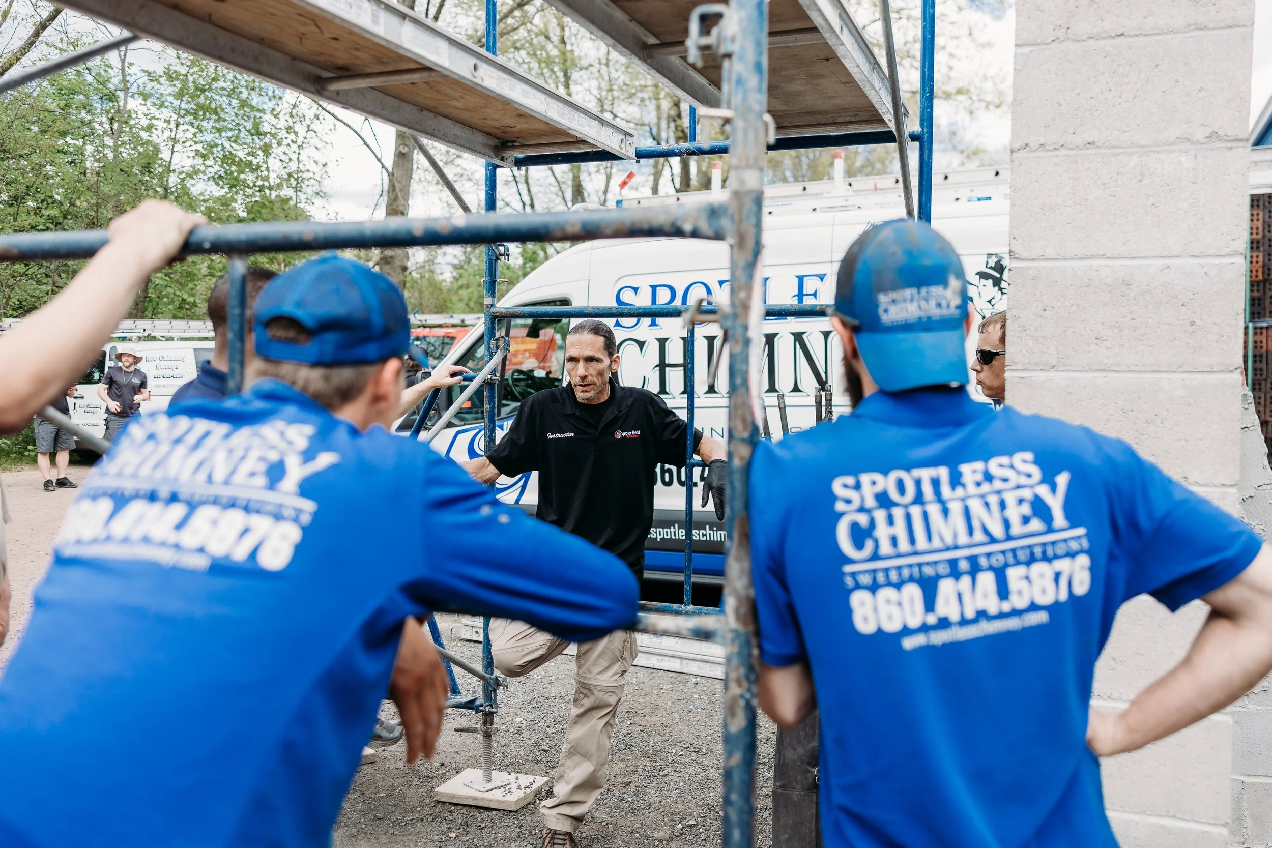 Workers in blue uniforms and caps discussing outside a construction site with scaffolding and a service van in the background.