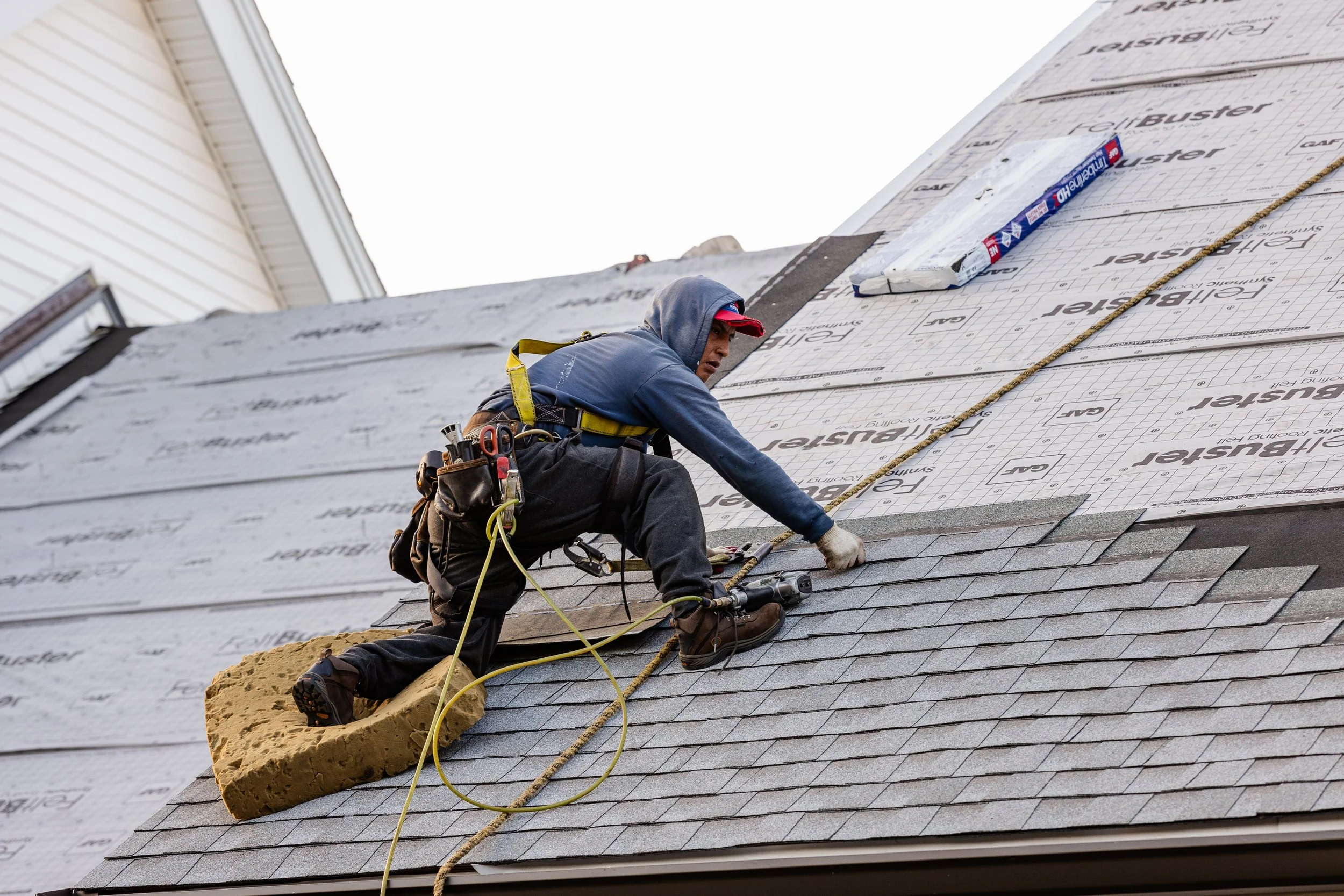A worker installing shingles on a roof, wearing safety gear including a harness and gloves.