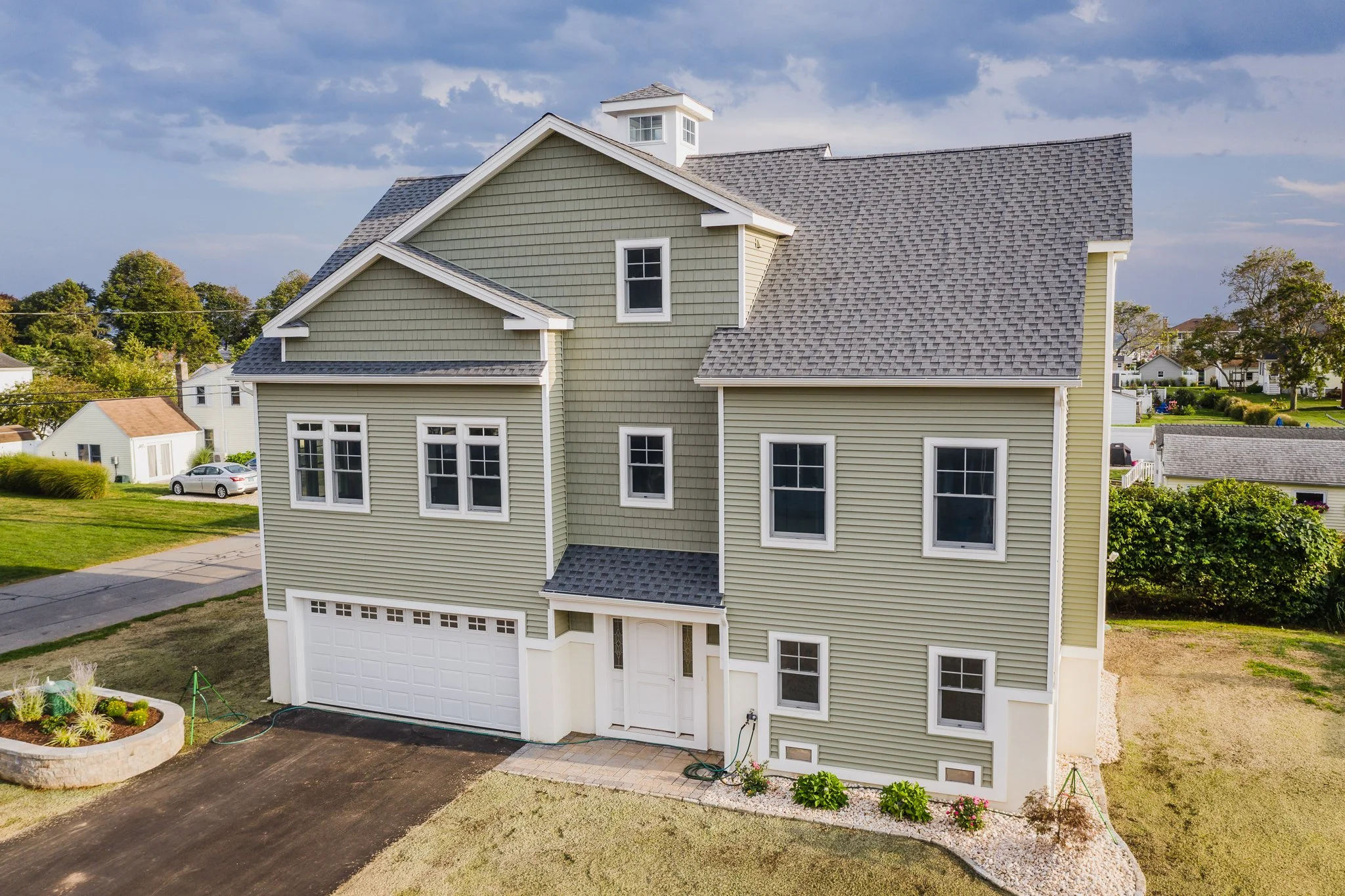 An aerial view of a multi-story, beige-colored house with a gray shingle roof in a suburban neighborhood, surrounded by green lawn and trees.