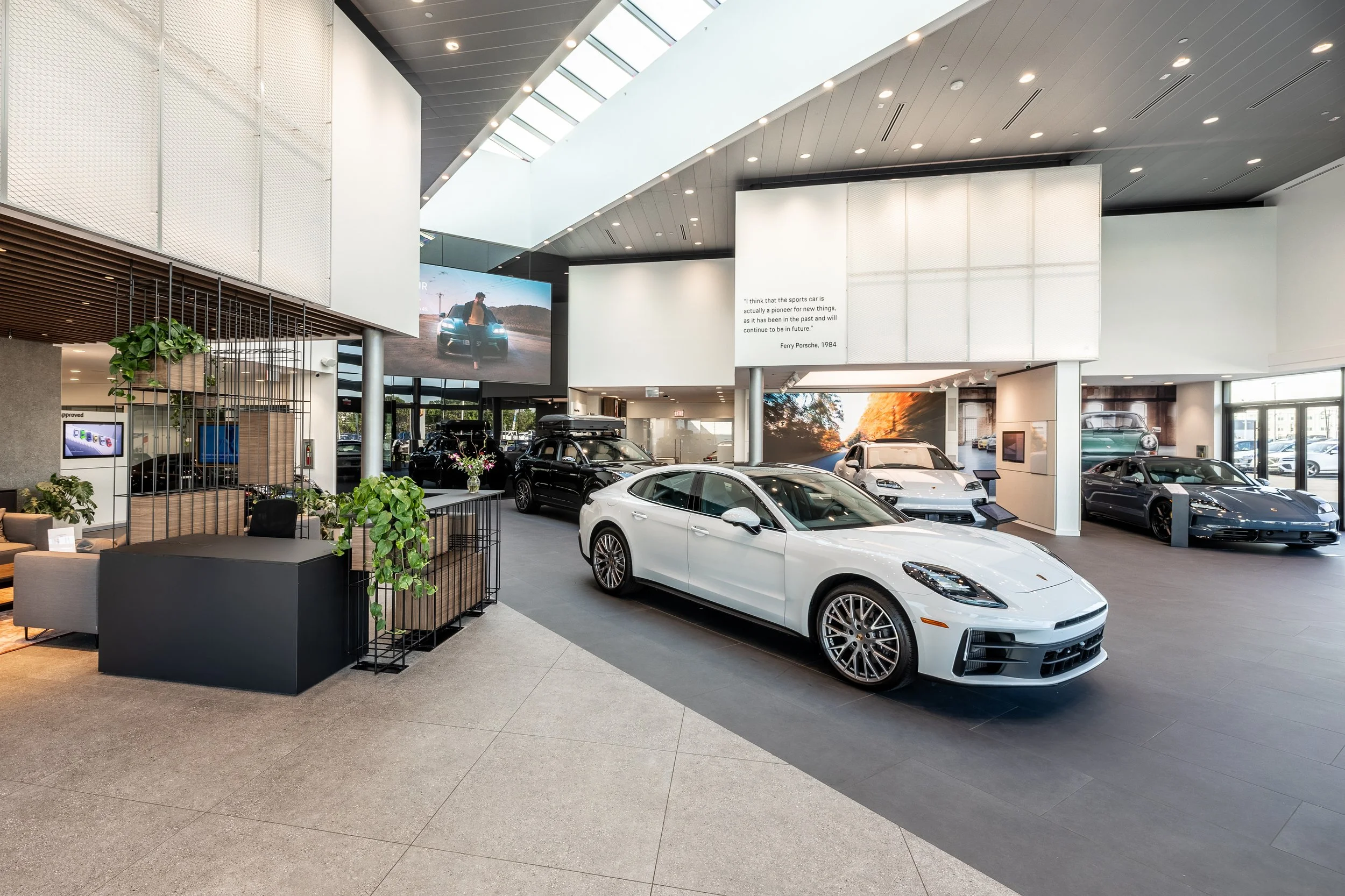 Inside a luxury car dealership showroom with multiple high-end cars on display, including a white sports car in the foreground, black SUVs, and a gray coupe, with modern interior design, large screens, and natural lighting.