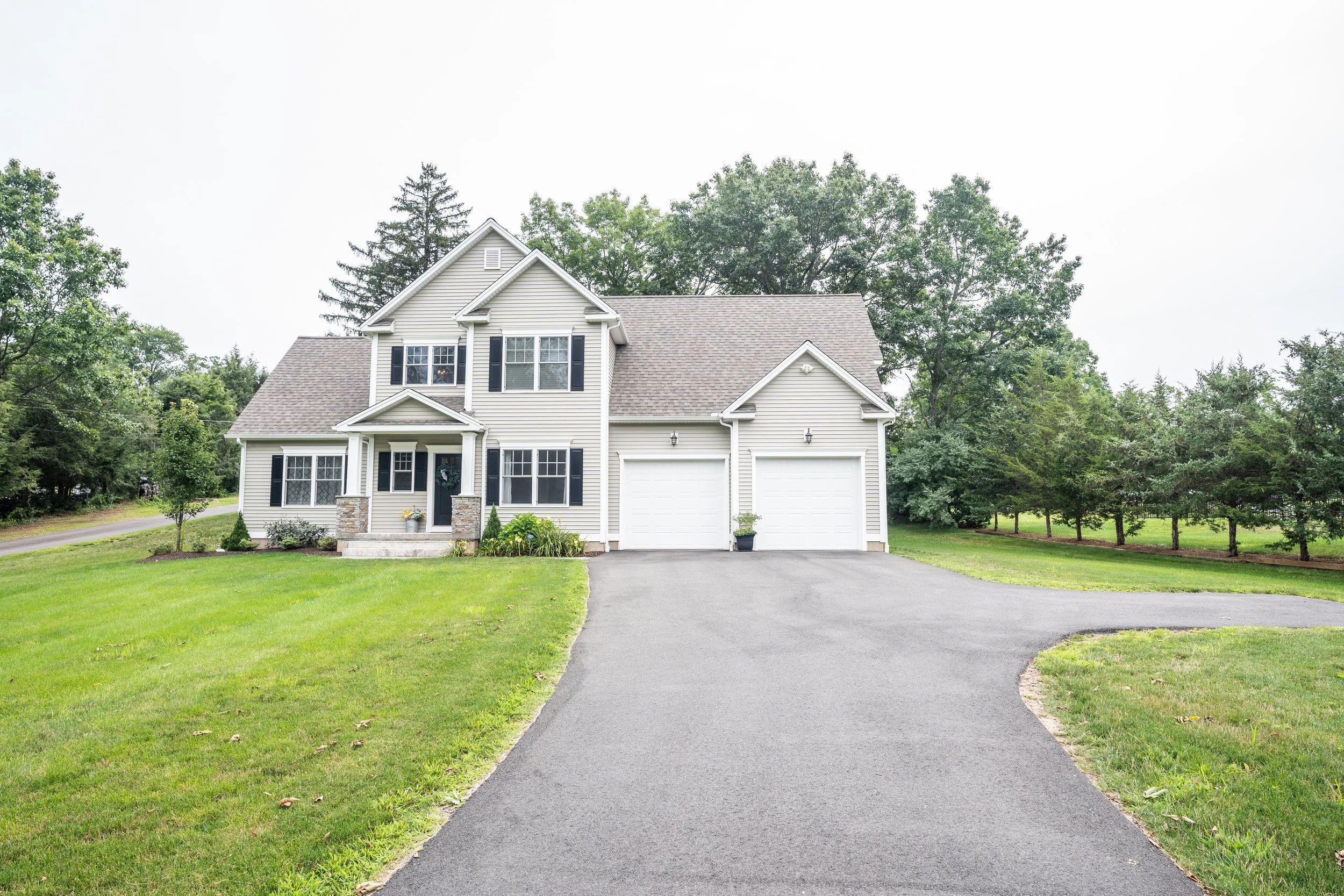 Front view of a white two-story house with black shutters, a two-car garage, and a paved driveway, surrounded by green lawn and trees.