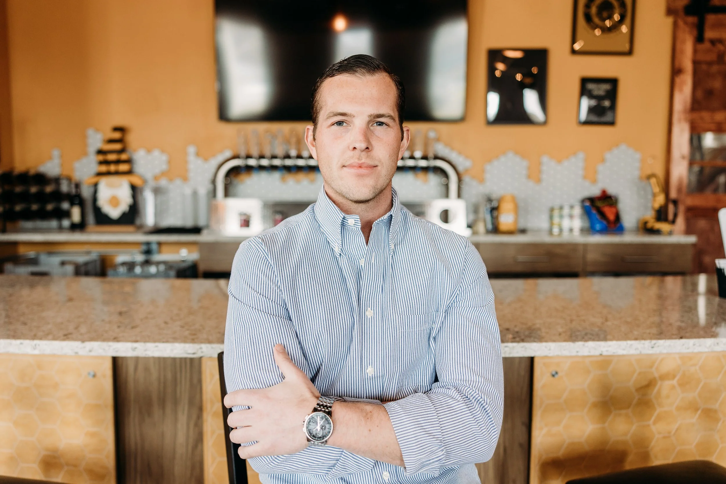 A young man with short dark hair, wearing a blue and white striped button-up shirt, standing with crossed arms in a cafe or restaurant. The background features a television, framed pictures, and a counter with various items, including bottles and too