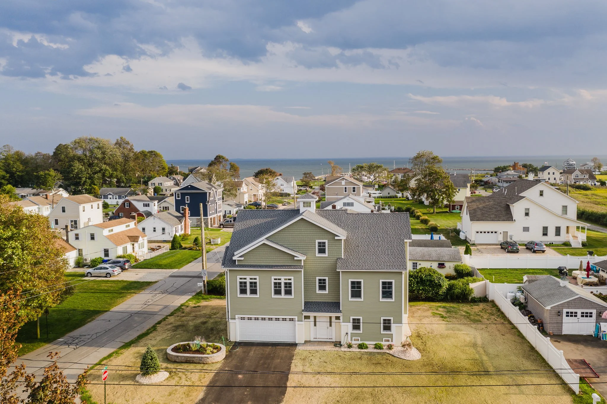 Aerial view of a suburban neighborhood near the coast with multiple houses, including a prominent green house in the foreground, green lawns, trees, and the ocean in the background under partly cloudy skies.