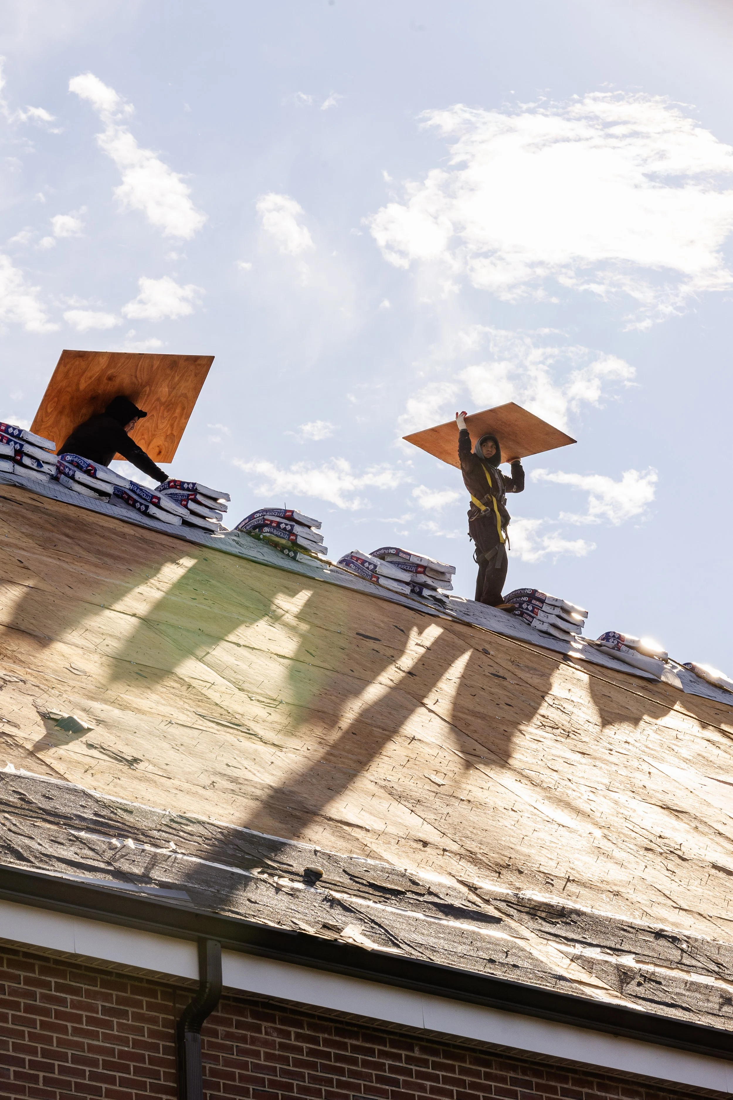 Two workers on a roof, carrying plywood sheets, with scaffolding and construction materials nearby, under a blue sky with clouds.