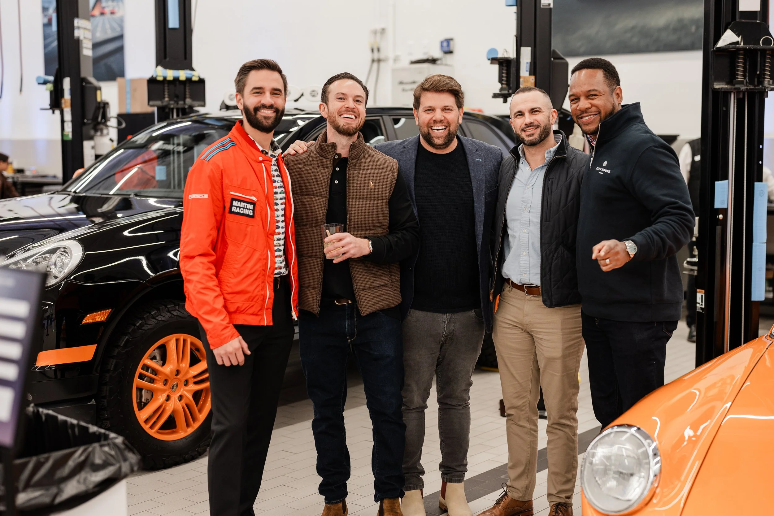 Five men standing together indoors next to a black car with orange wheels, smiling and posing for a photo, in what appears to be a car dealership or showroom.