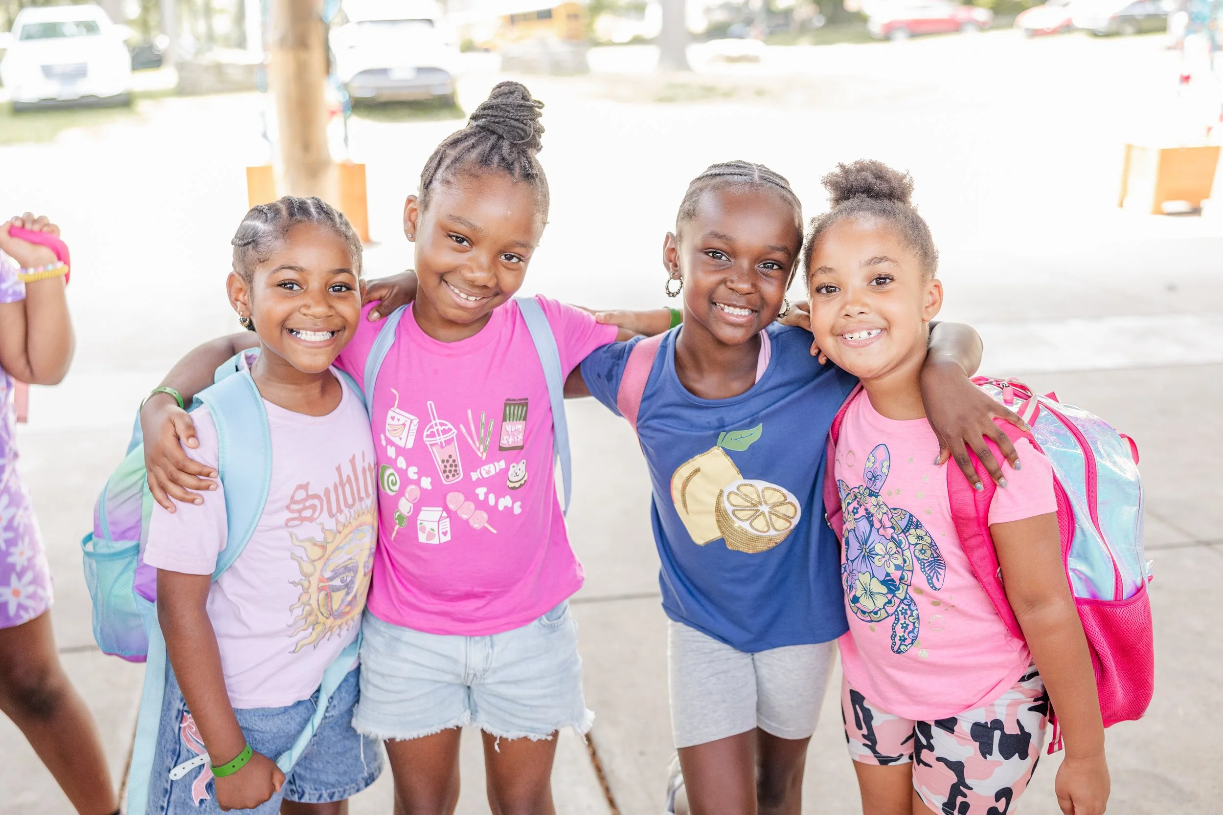 Four young girls standing close together with arms around each other, smiling at the camera during the day outdoors, with a background of parked cars and a wooden support beam.