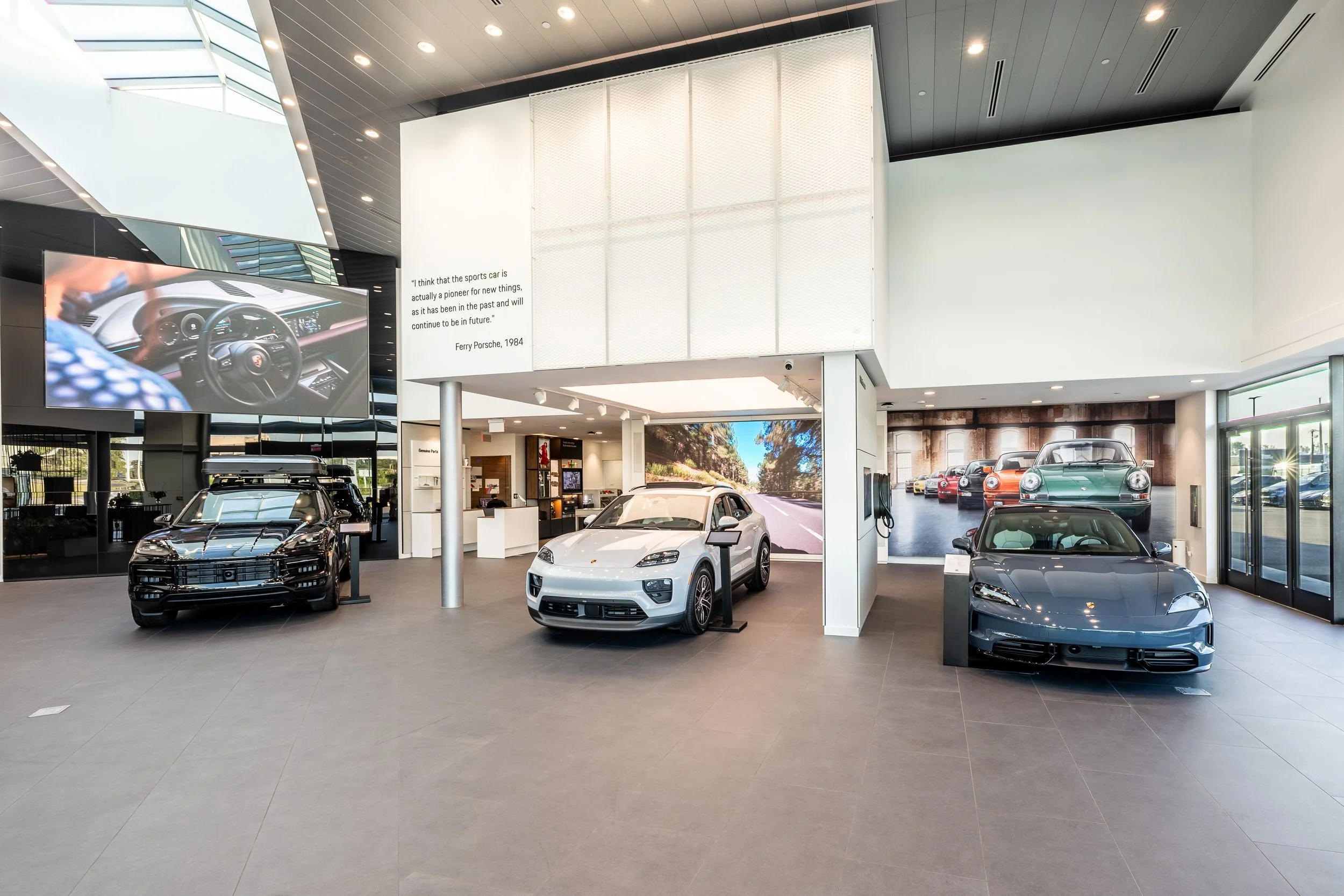 Inside a car dealership showroom displaying several luxury sports cars, including a black, white, and gray car, with a large digital screen showing a car interior and a quote about sports cars.