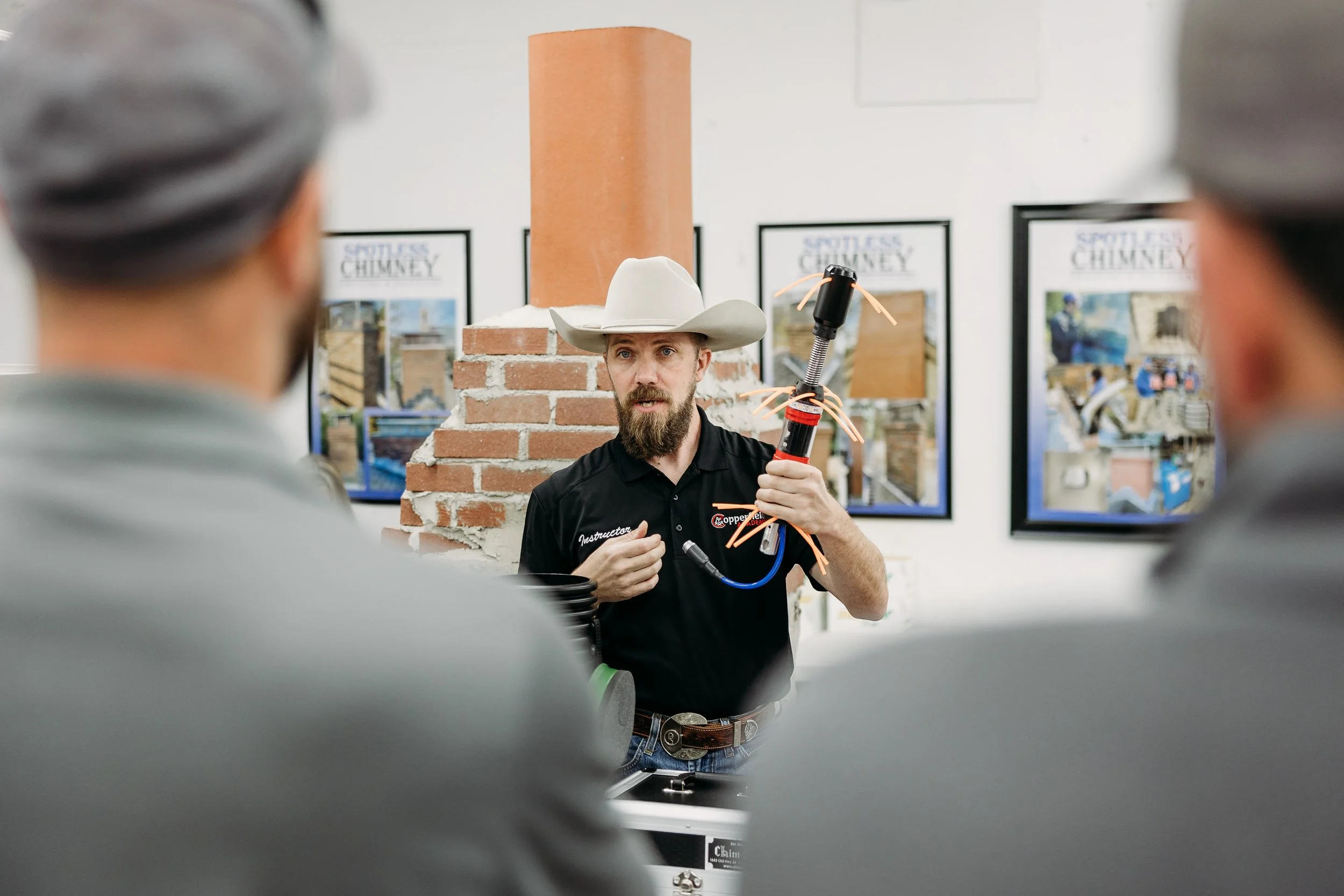 A man with a white cowboy hat explaining a welding torch to two people in front of him during a demonstration in a room with brick and wall posters.