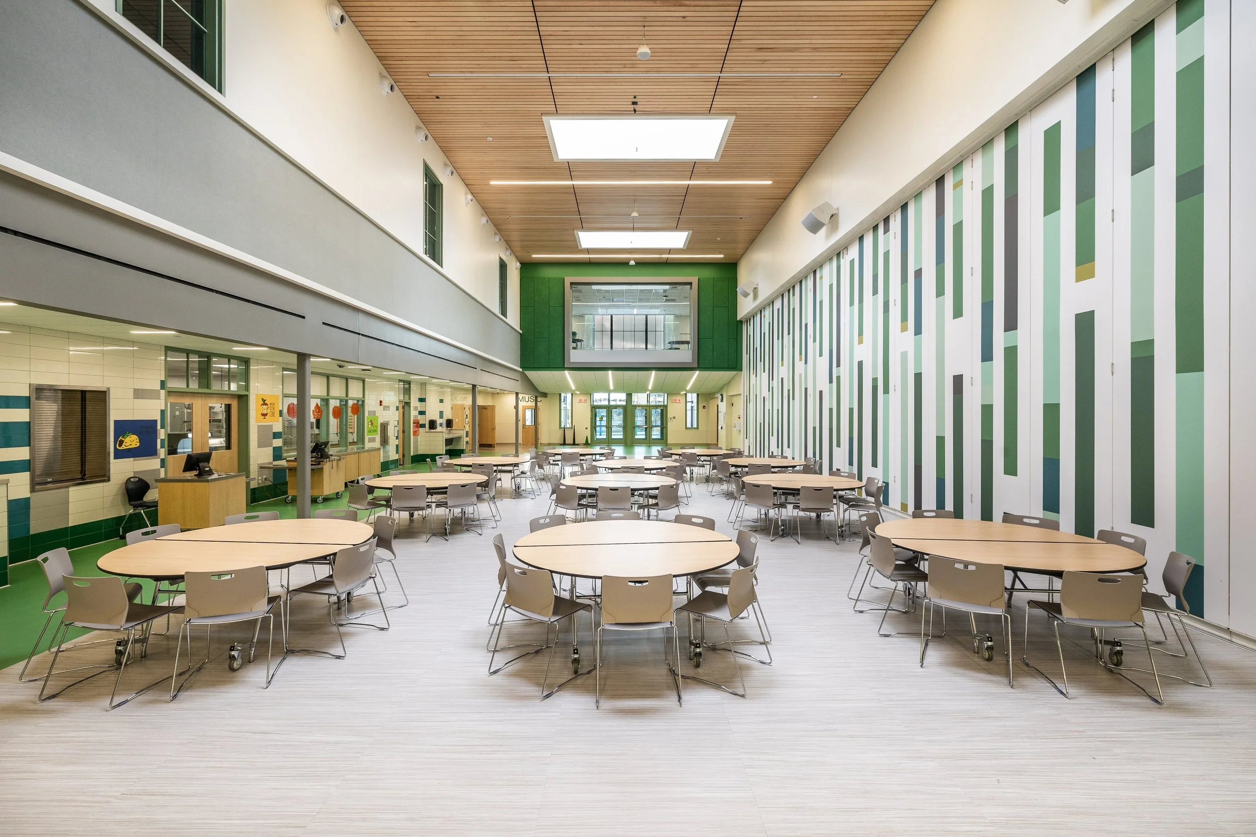 A spacious modern school cafeteria with round tables, gray chairs, and a colorful striped wall on the right. The ceiling has wooden panels with bright square lights, and the back wall features large glass windows and doors.