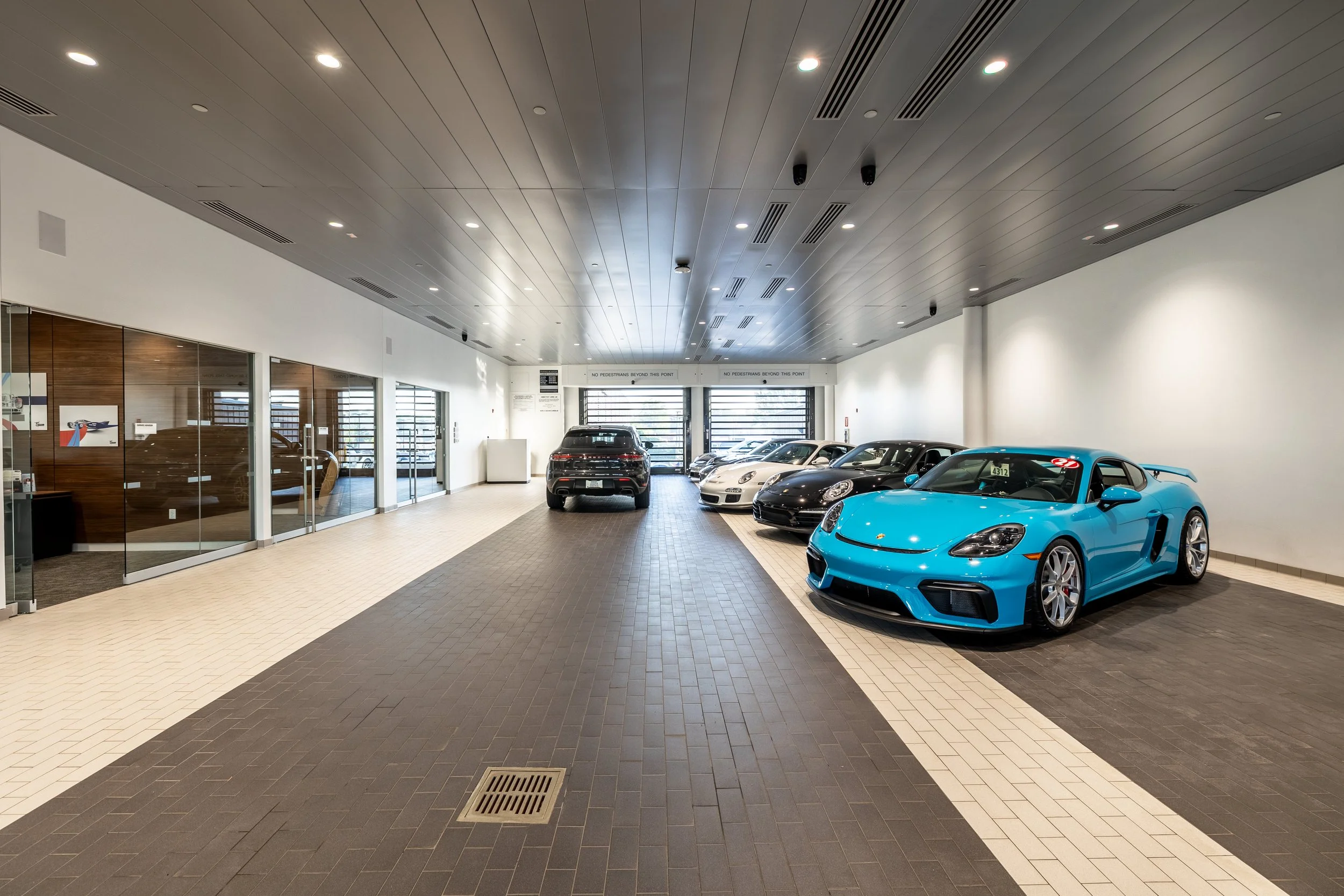 Indoor car dealership with several sports cars, including a bright blue Porsche in the foreground, and other luxury vehicles parked behind it, with a modern, well-lit interior with gray and beige tile flooring.