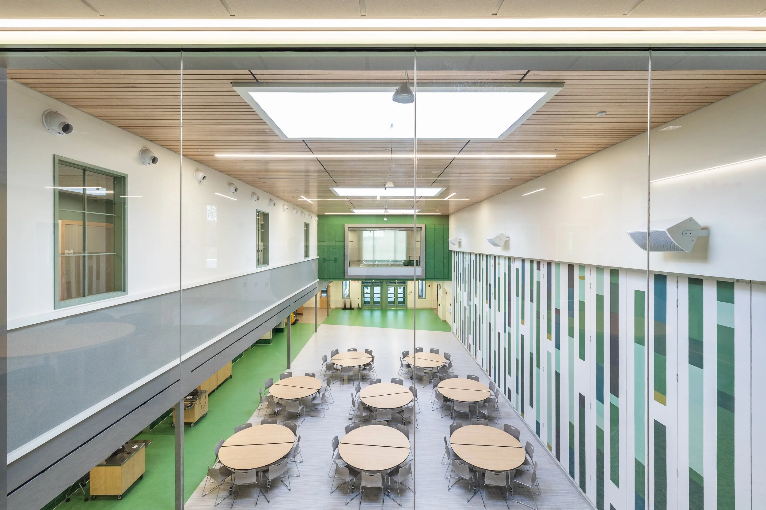 Interior of a modern cafeteria with round tables and chairs, a green accent wall, windows, and a wooden ceiling with skylights.