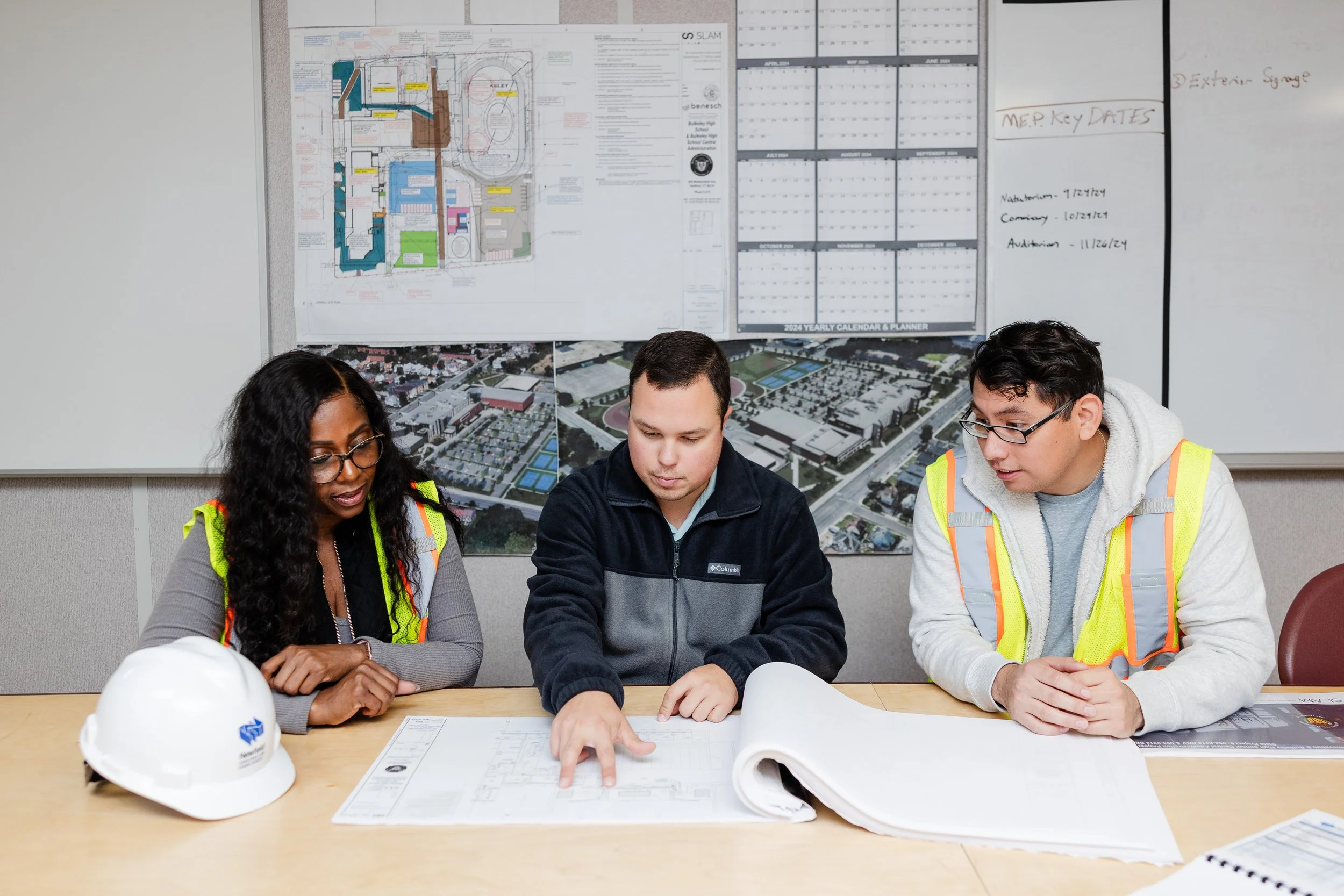 Three construction professionals in safety vests reviewing blueprints and plans at a table, with architectural maps and aerial site images on the wall behind them.