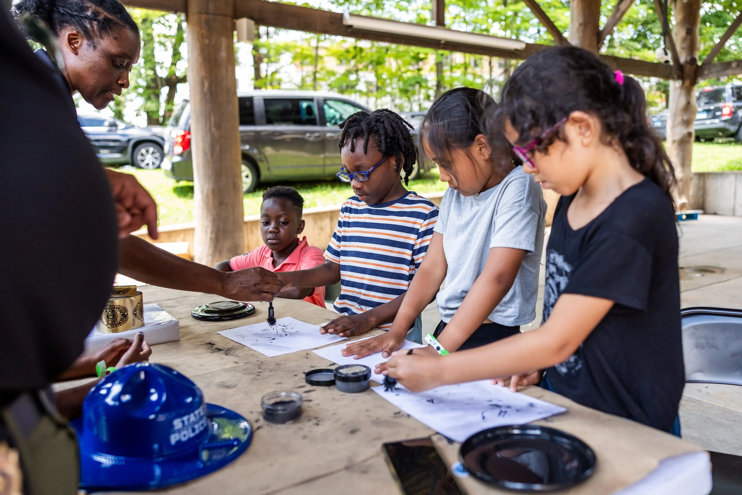 A group of children and an adult participating in an outdoor educational activity at a table, under a wooden canopy, with cars and trees in the background.