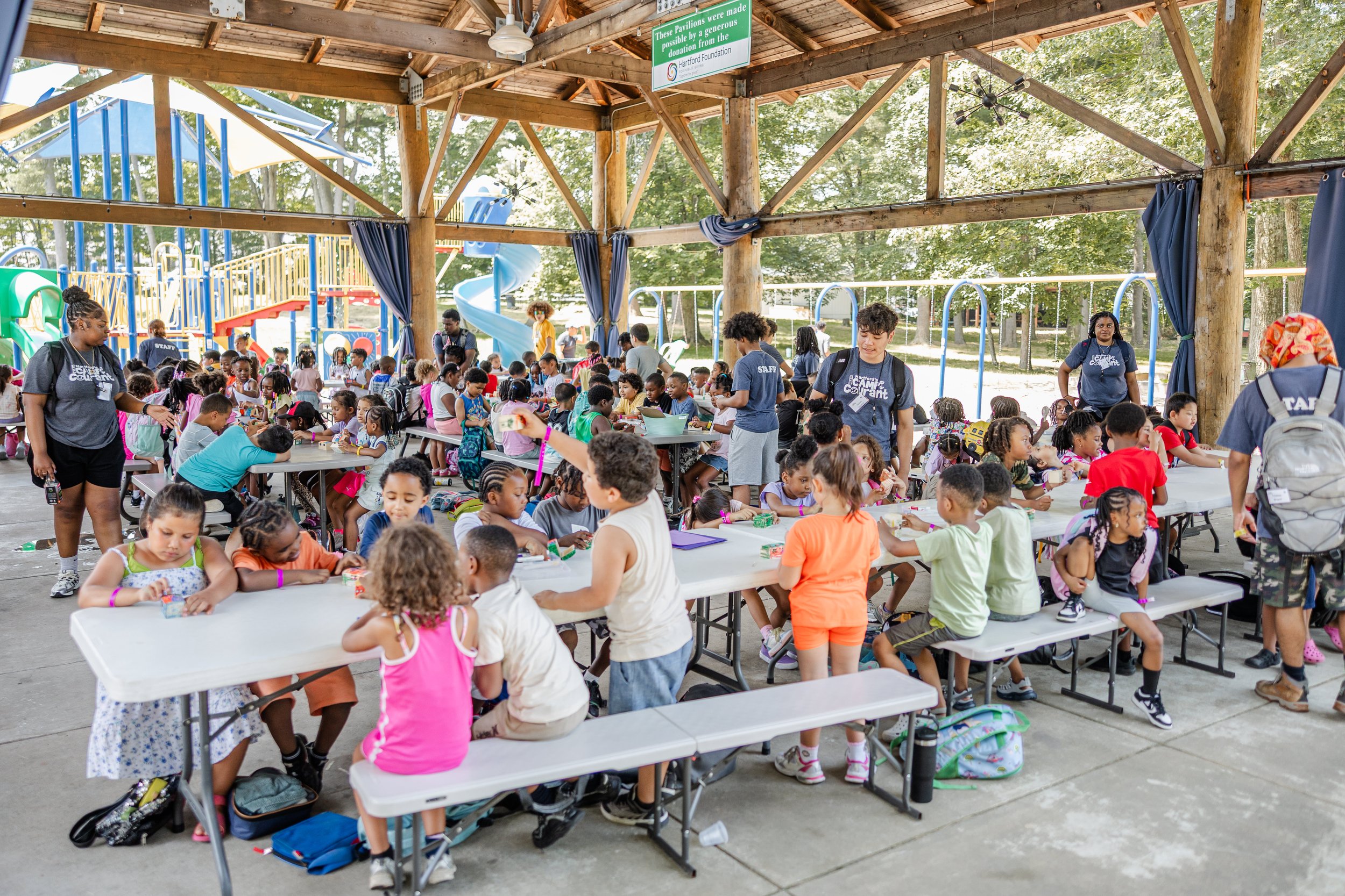 Children participating in a camp activity under a large wooden pavilion with picnic tables, surrounded by playground equipment and trees.
