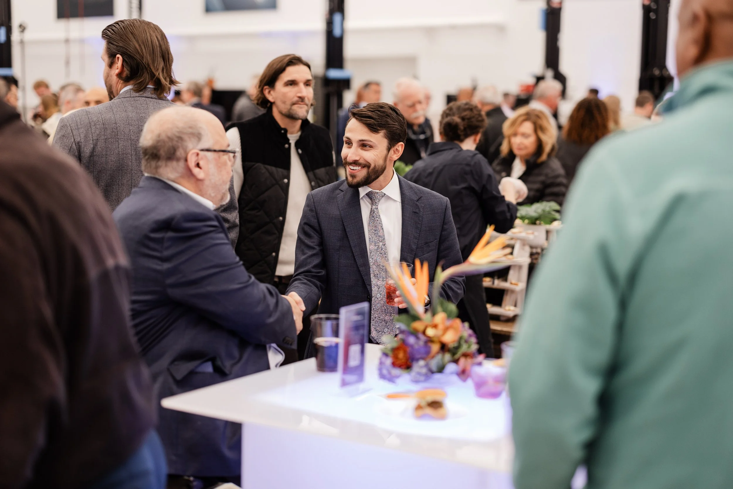 A man in a dark suit shakes hands with an older man in a navy blazer at a busy indoor event, with a floral centerpiece on the table they are standing at.