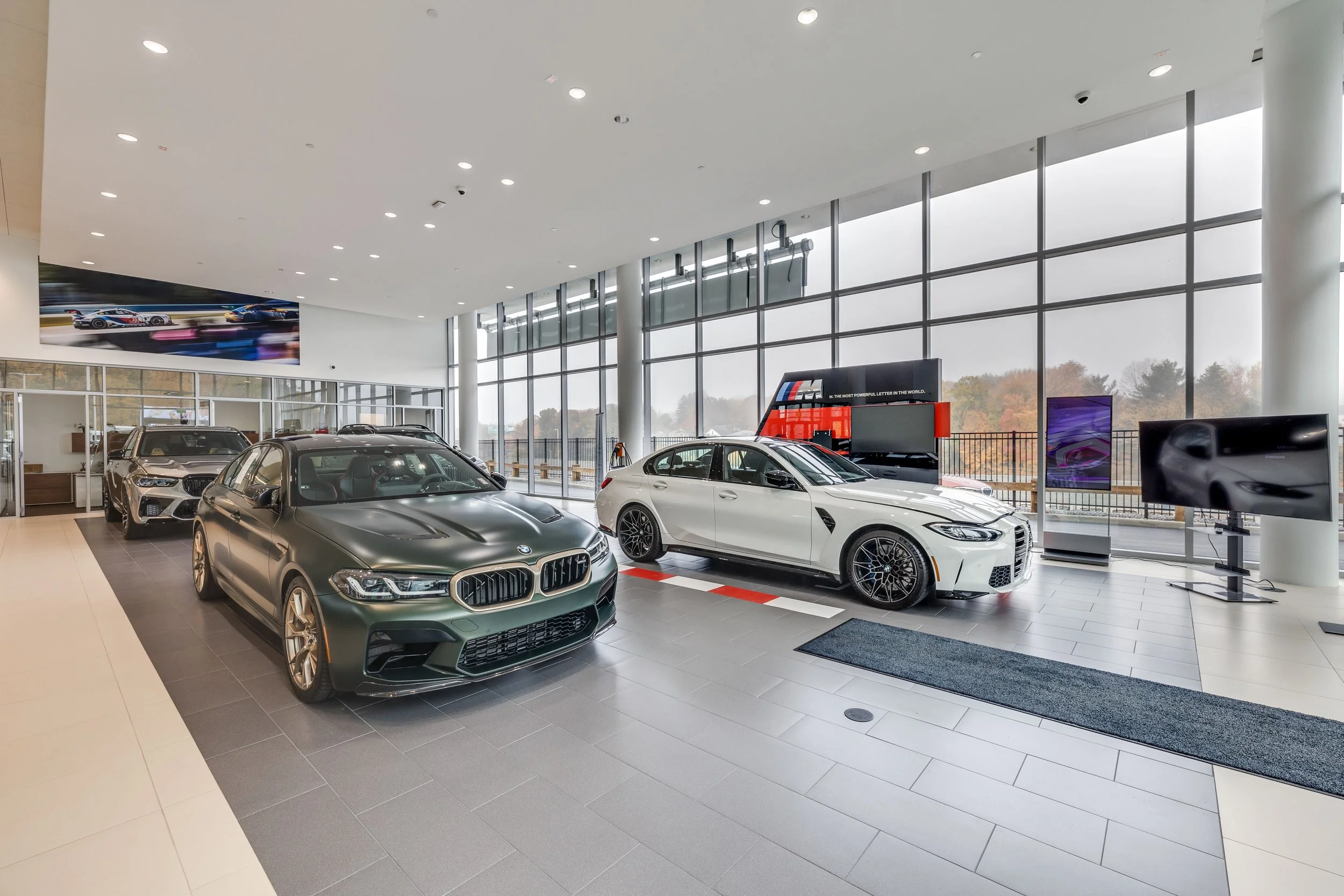 Showroom with several BMW and other luxury cars displayed, large windows, and digital screens.
