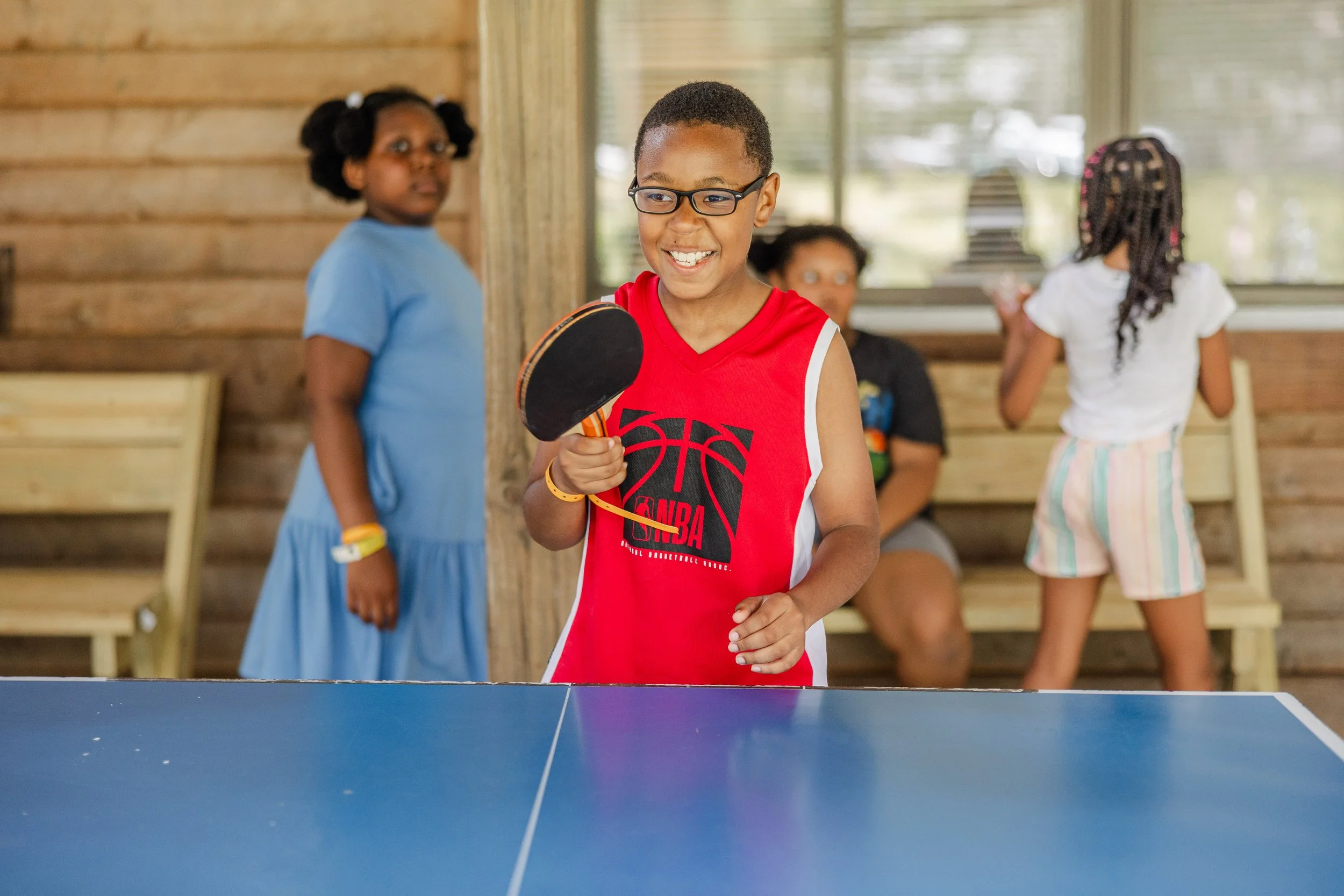 A group of children playing indoor table tennis. One boy in a red basketball jersey is holding a ping pong paddle and smiling, while three girls and a woman are in the background, some standing and some sitting, inside a wooden-paneled room with larg