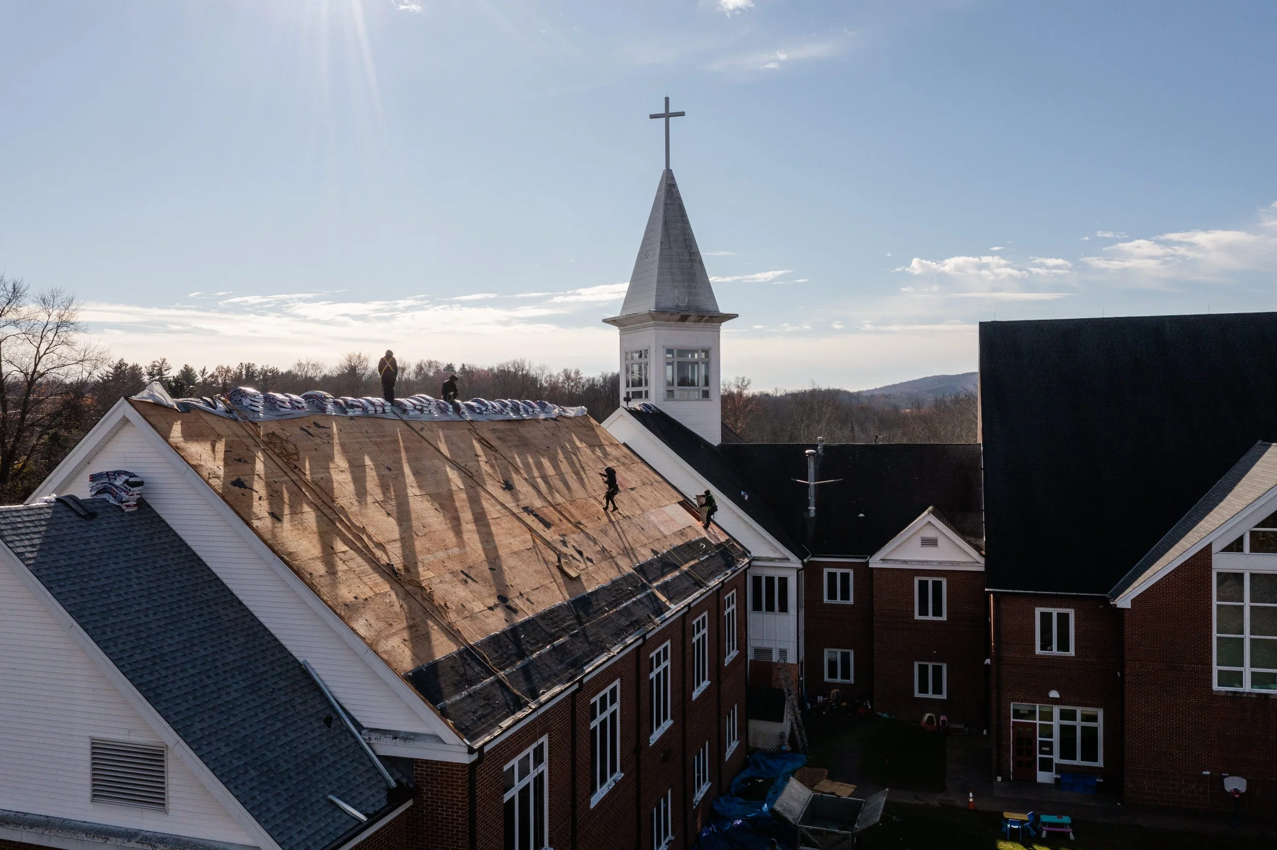 Church building under roof renovation, workers on the roof removing shingles, church steeple with cross in the background, clear sky with the sun shining.