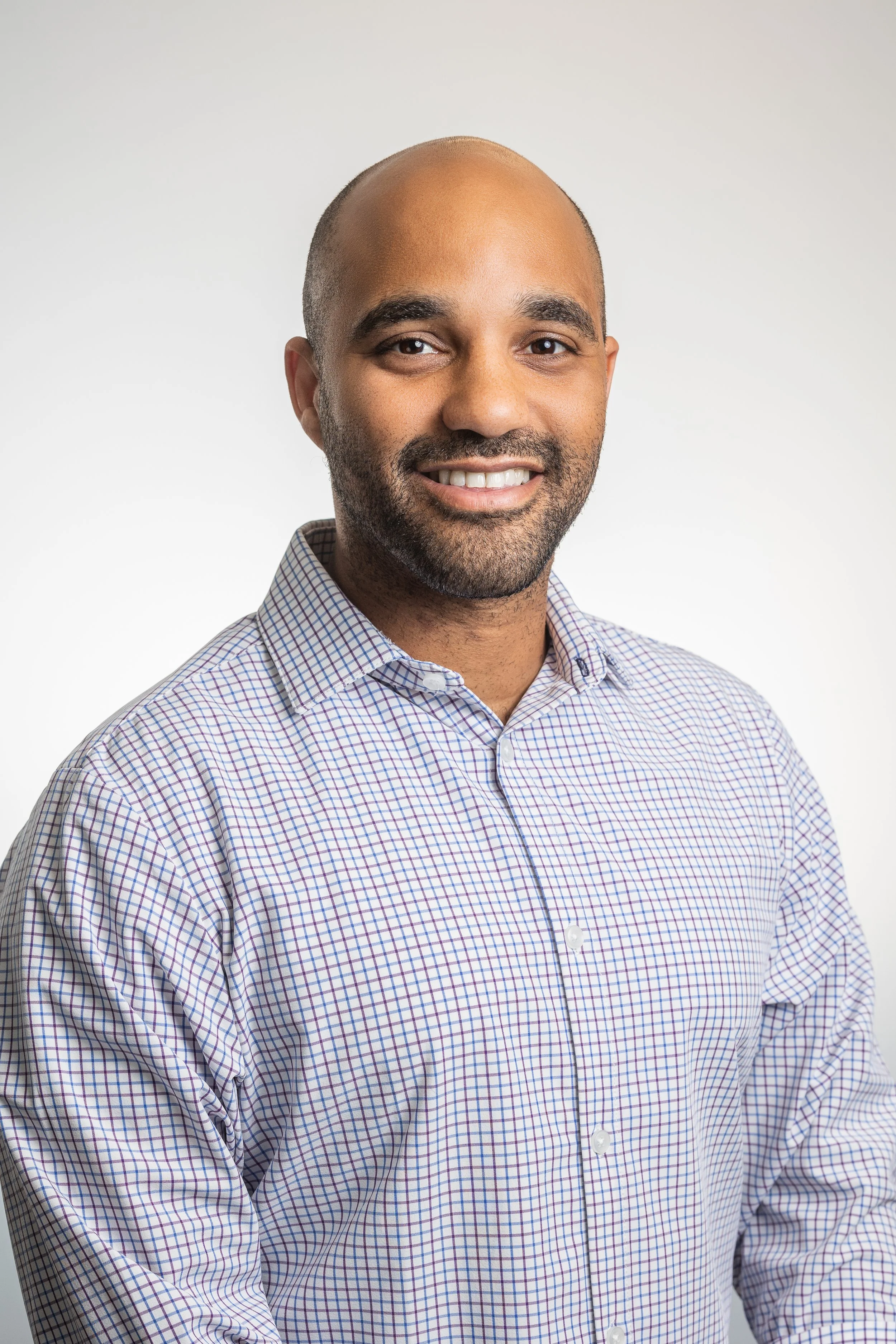 Headshot of a smiling man with a beard and a shaved head, wearing a white and purple checkered button-up shirt, against a plain white background.