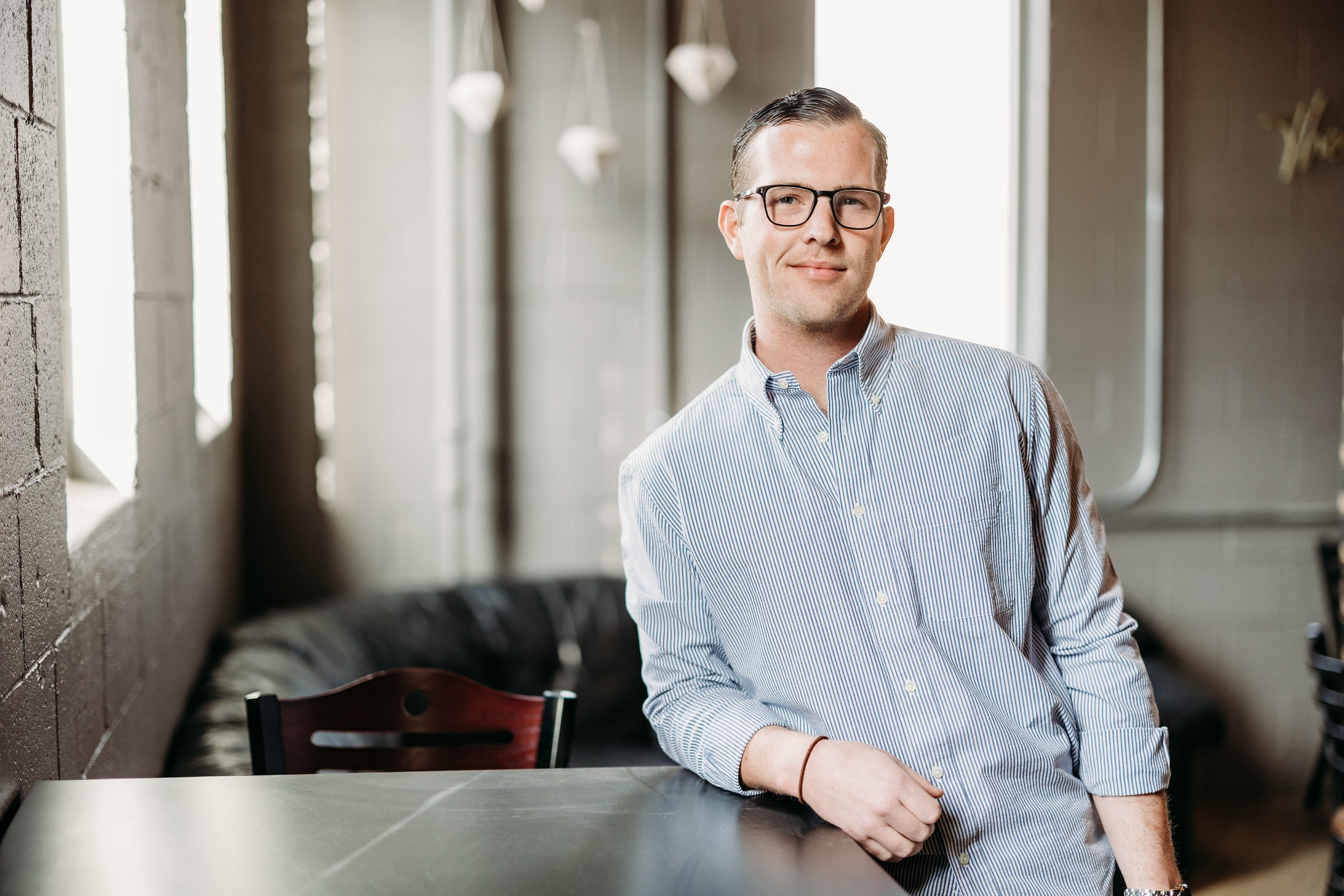 A man with glasses in a striped shirt leaning on a table in a modern, well-lit room.