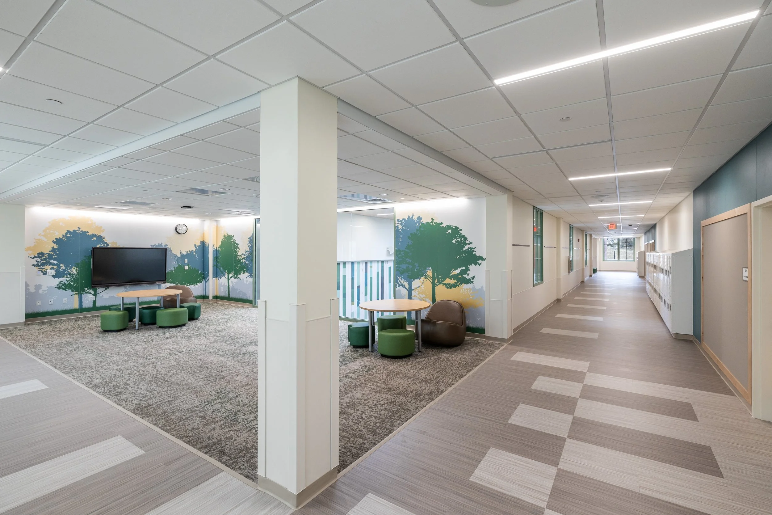 A bright, modern school hallway with lockers on the right side and a small seating area with a mural of trees on the left. The ceiling has white panels with integrated lighting, and the floor features a pattern of light and dark grey tiles.
