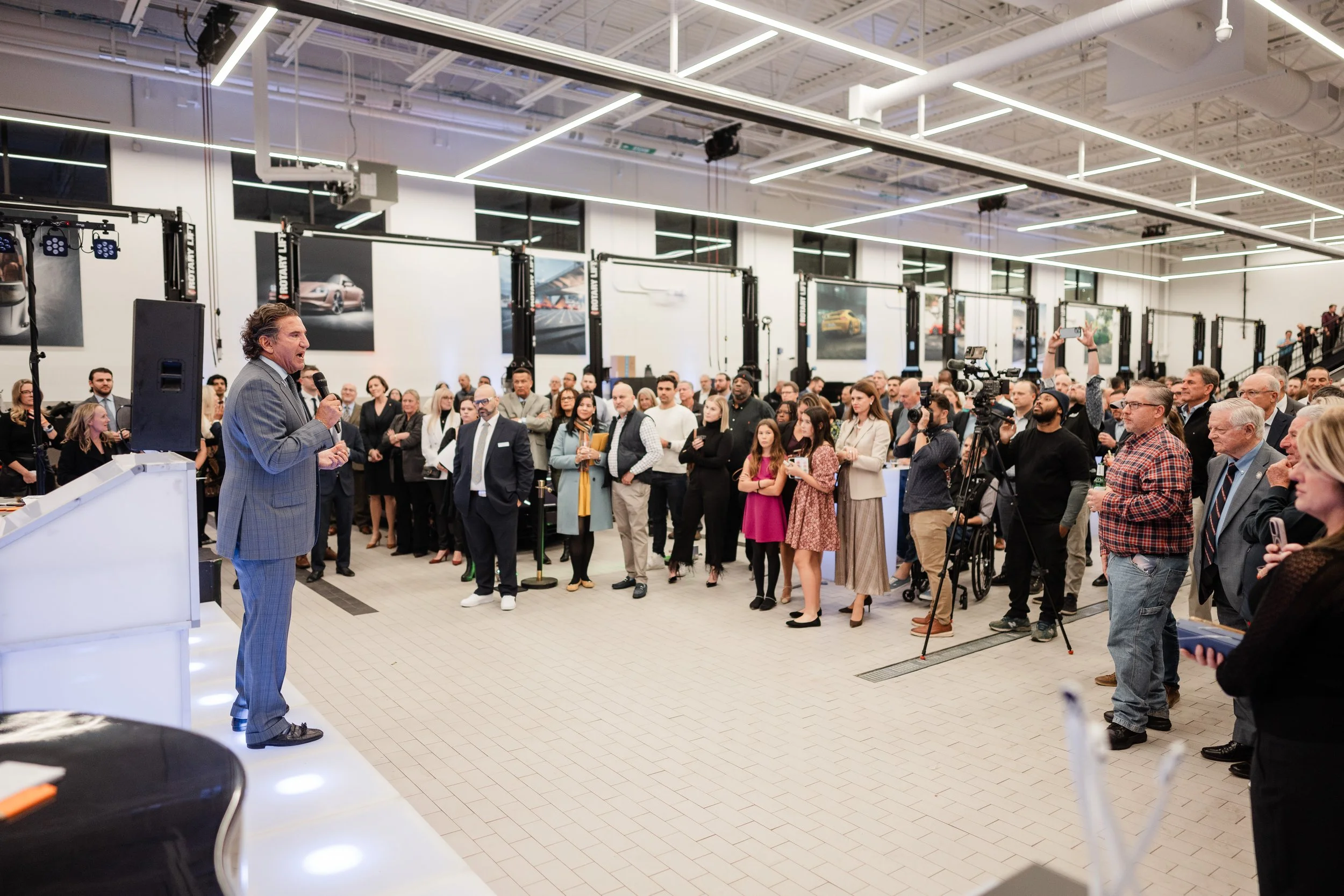 A man in a gray suit speaks into a microphone on a stage surrounded by an audience in a large, modern showroom with car posters on the walls.