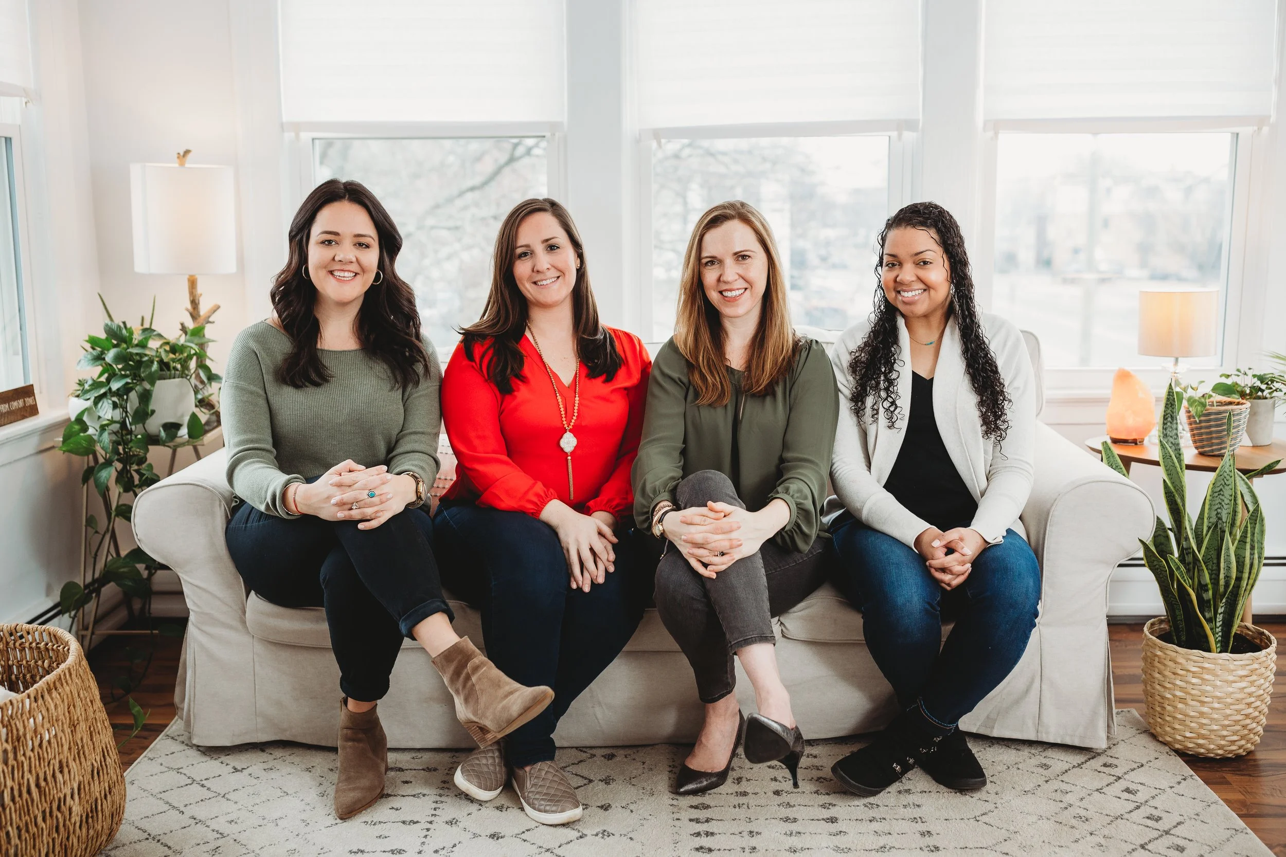 Four women sitting on a light-colored sofa in a bright room with large windows, potted plants, and lamps.