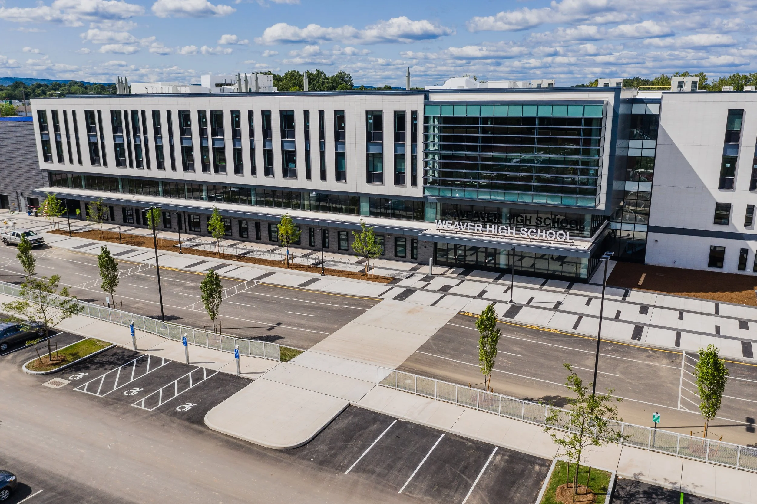 A modern school building with a large glass entrance, surrounded by a parking lot with marked spaces, some trees, and a sidewalk, under a partly cloudy sky.