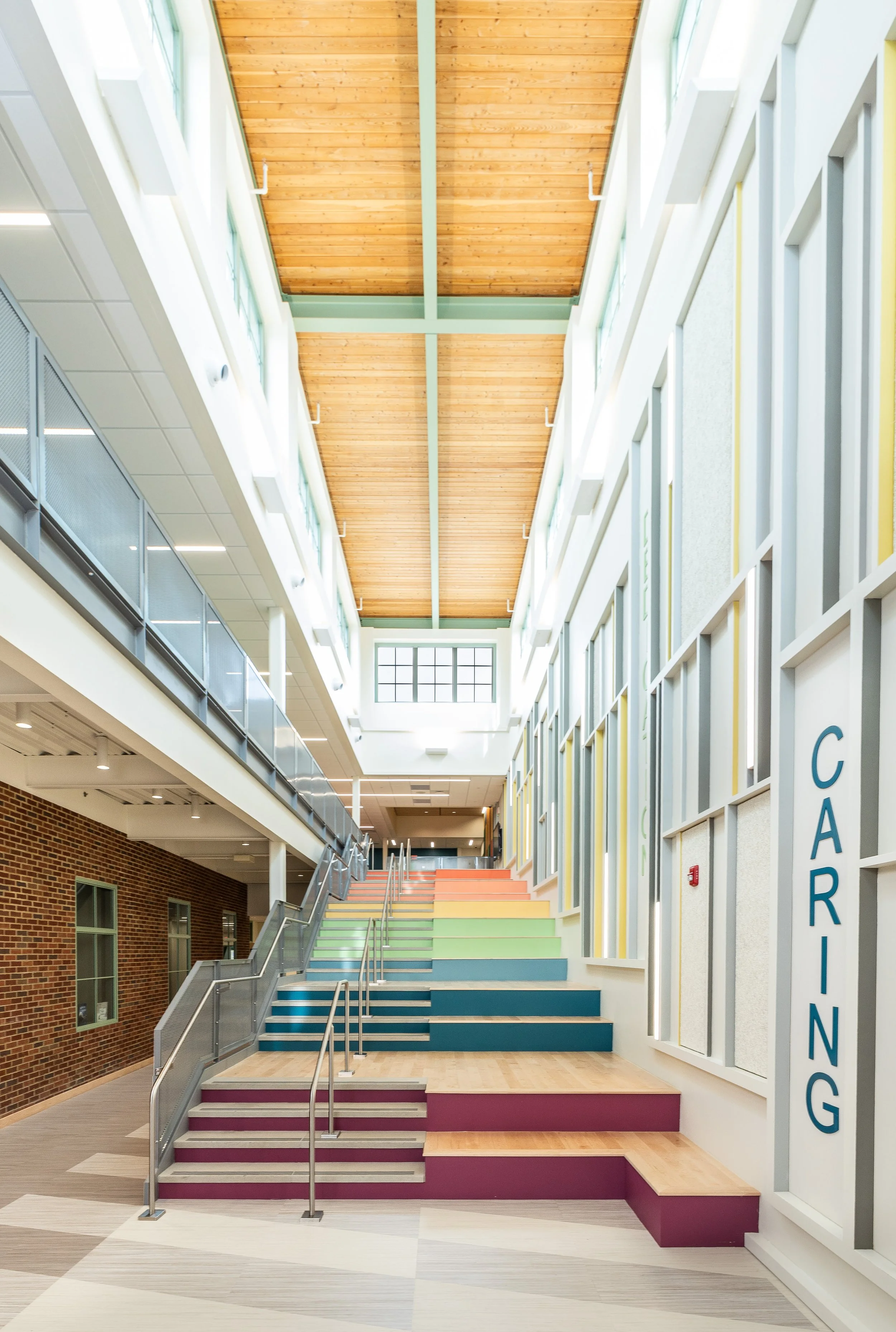 Interior of a modern building with colorful steps leading up to a second floor, featuring a high wooden ceiling, glass walls, and bright natural light.