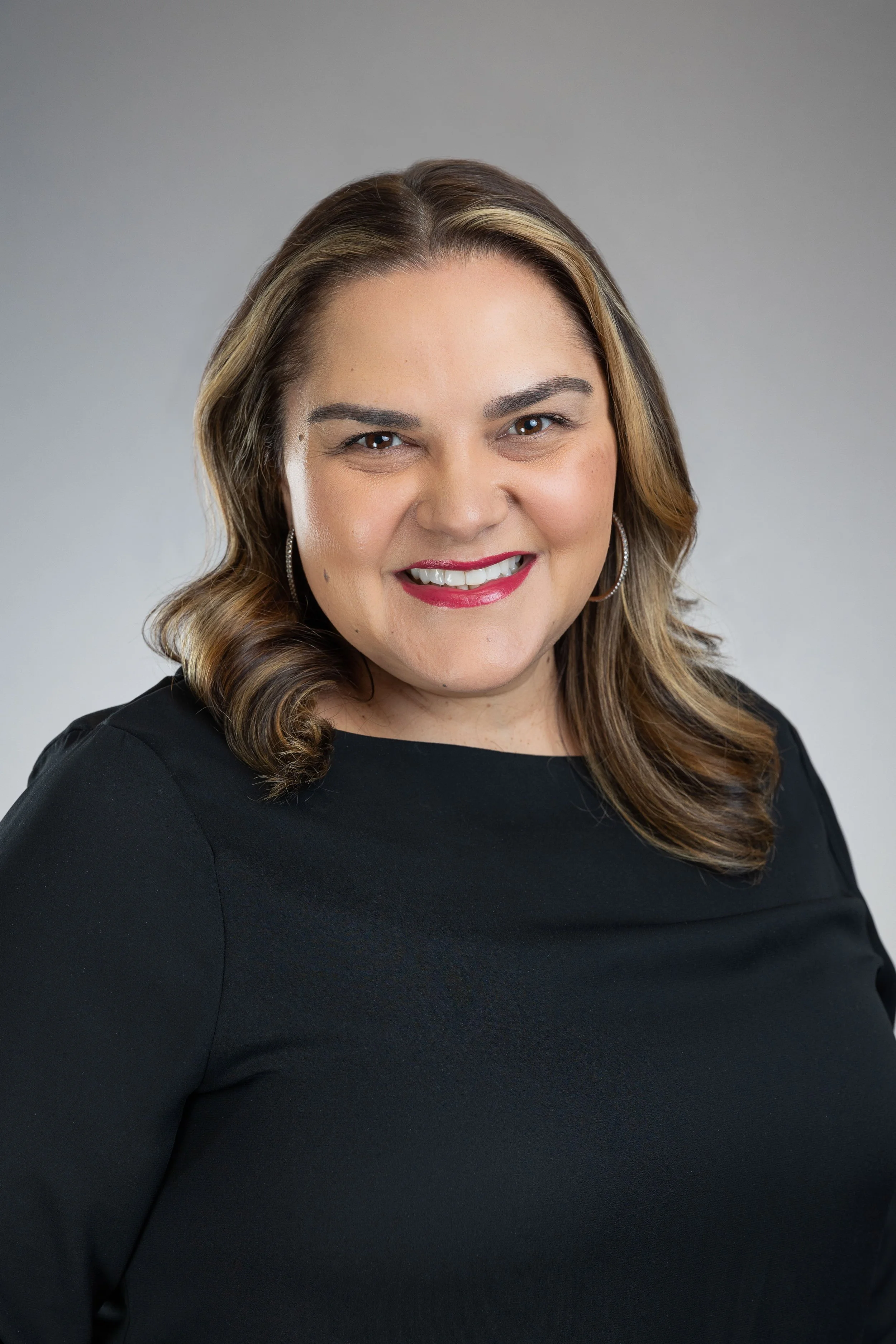 A woman with shoulder-length brown hair and blonde highlights smiling, wearing a black top and hoop earrings, against a plain grey background.