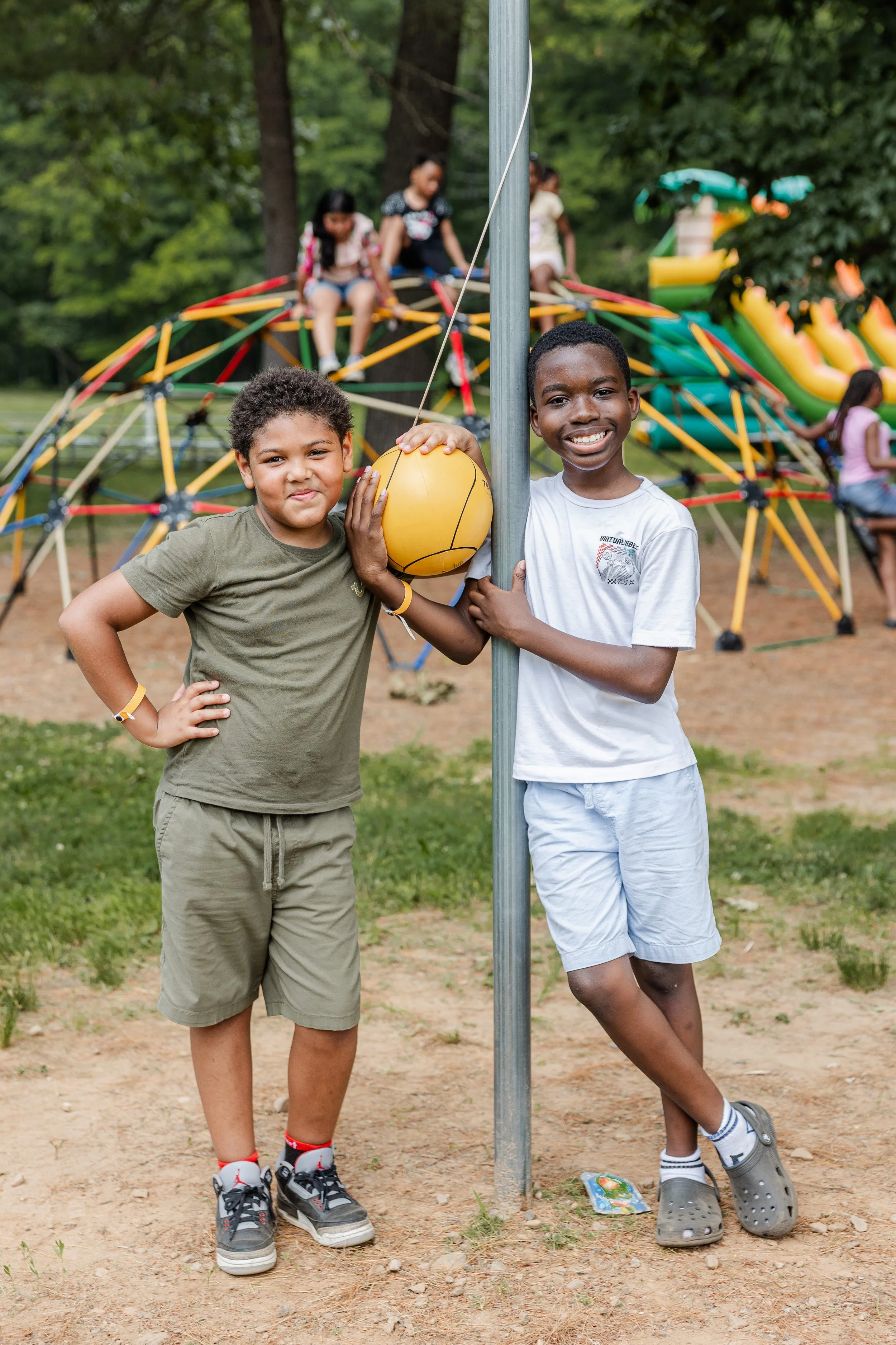 Two boys are at a playground, holding a yellow ball and standing on either side of a metal pole. Behind them, there is a colorful climbing structure with children playing on it and a slide. The scene is outdoors with green trees in the background.