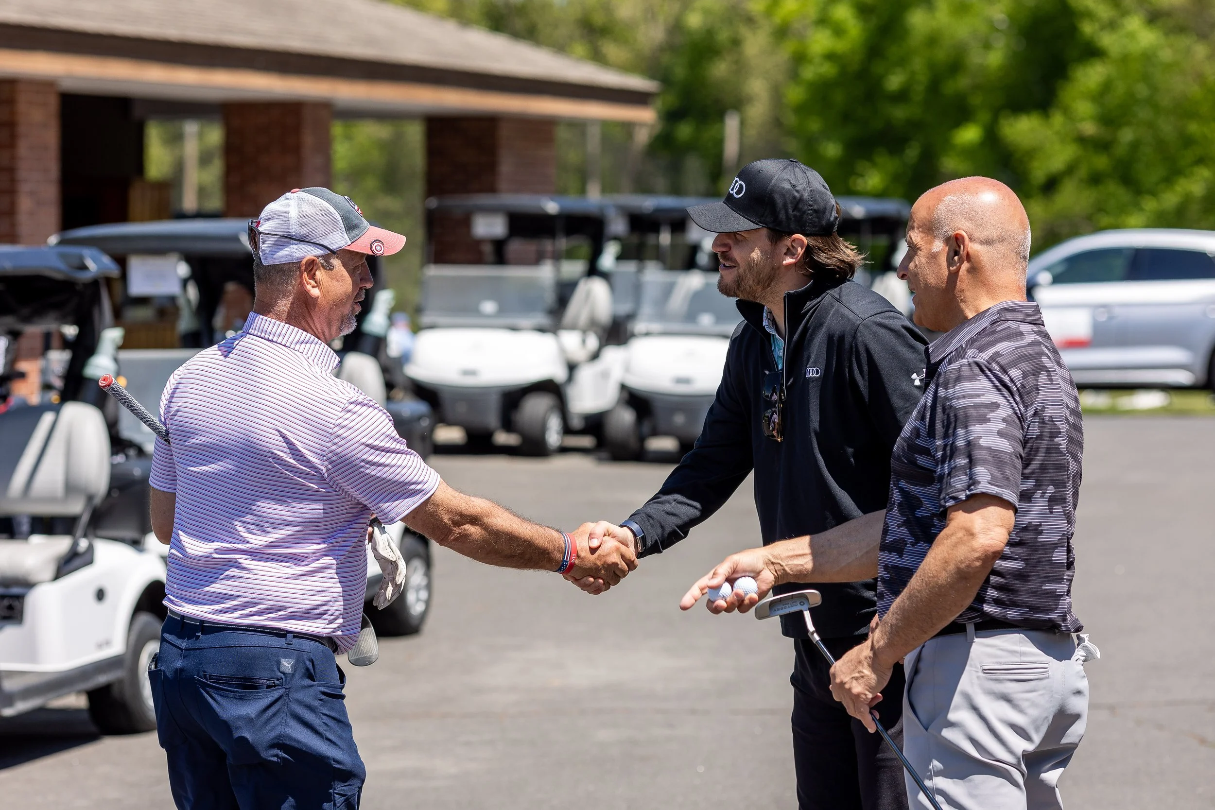 Three men on a golf course shaking hands, with golf carts and trees in the background.