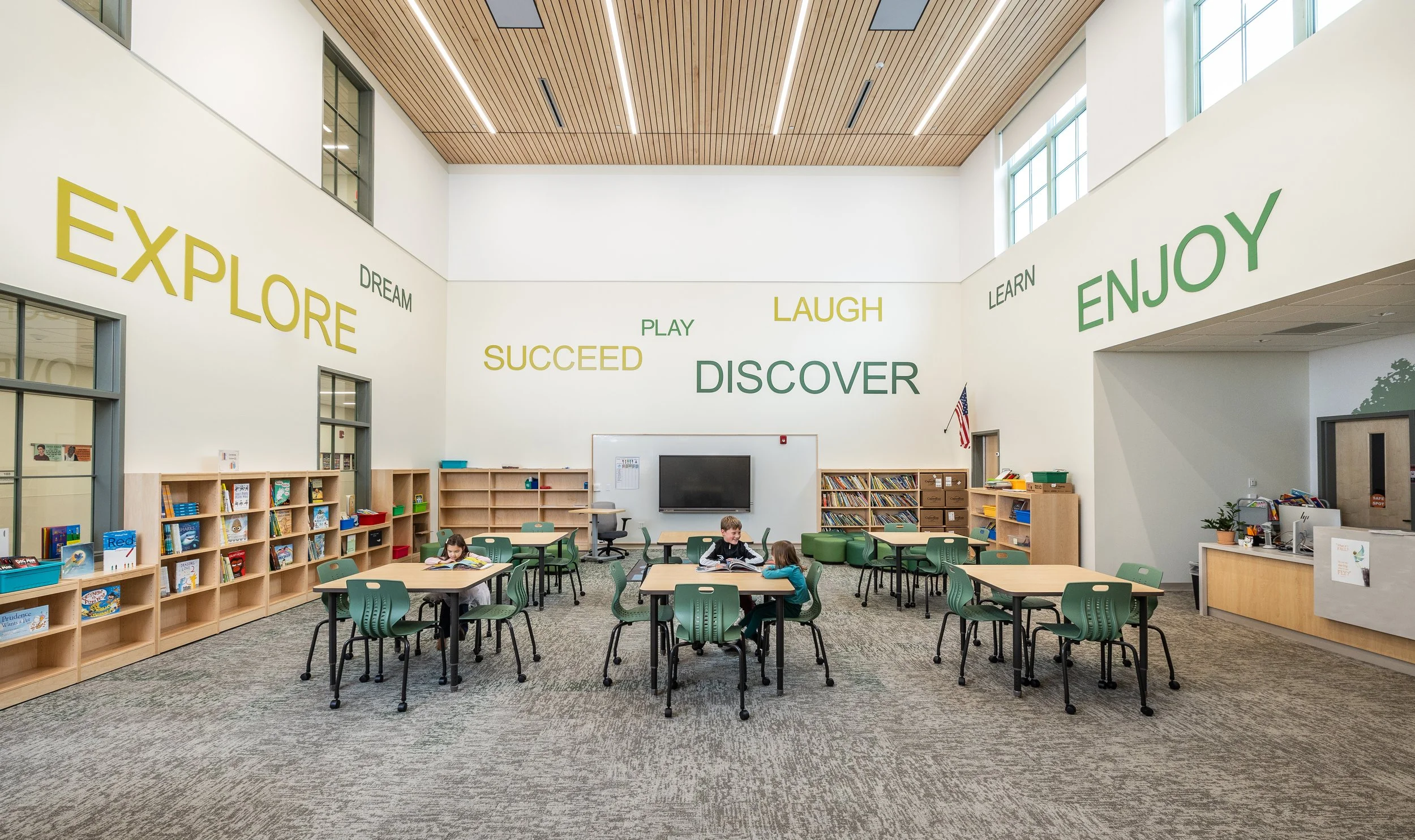 A brightly lit modern classroom with high ceilings, wooden slat ceiling, and walls decorated with words like Explore, Discover, Learn, and Enjoy in various colors. There are multiple tables with green chairs, children reading, bookshelves filled with