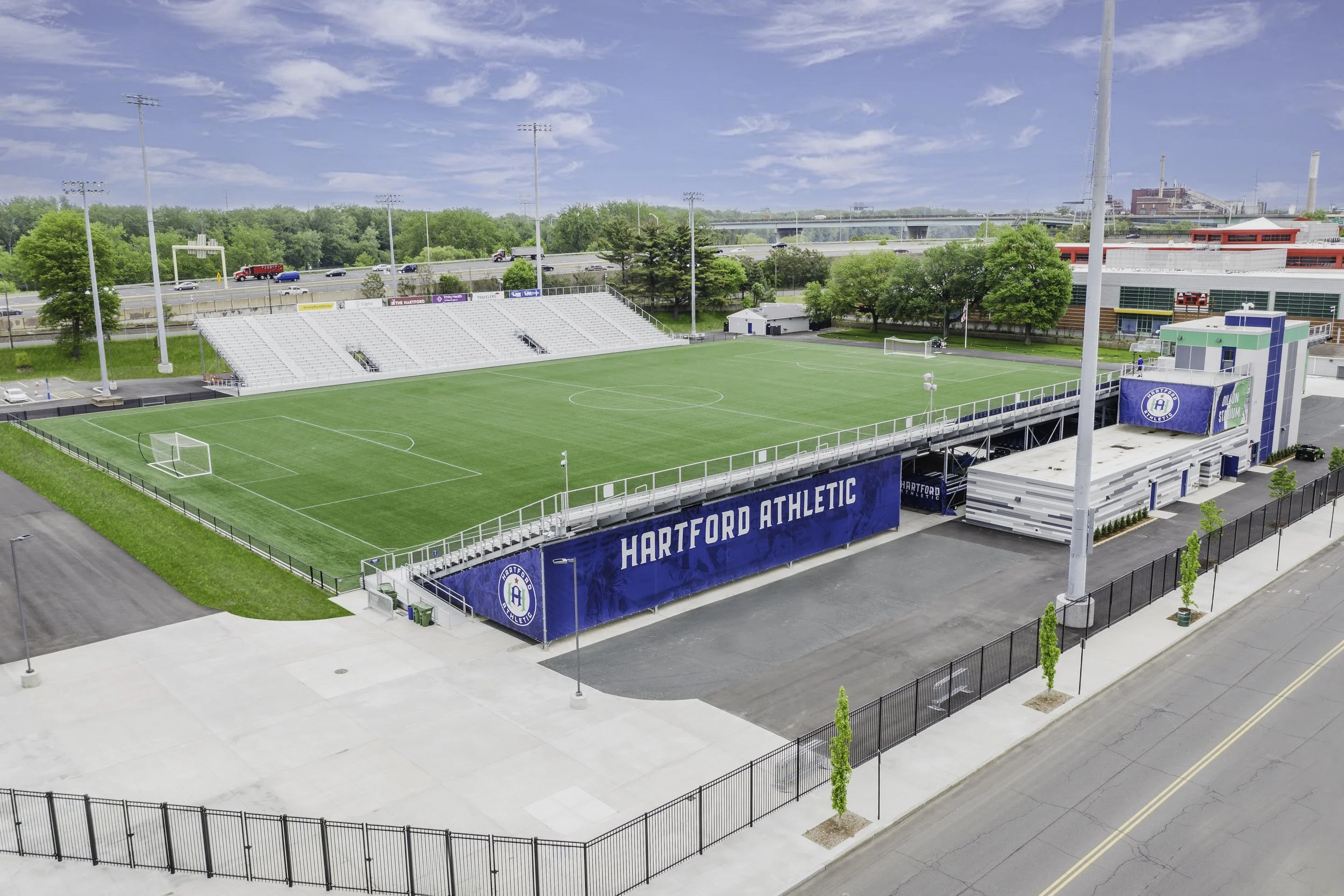 An outdoor soccer stadium with a green field, seating area, and blue Hartford Athletic banners, surrounded by a black fence, with a parking lot and trees in the background.
