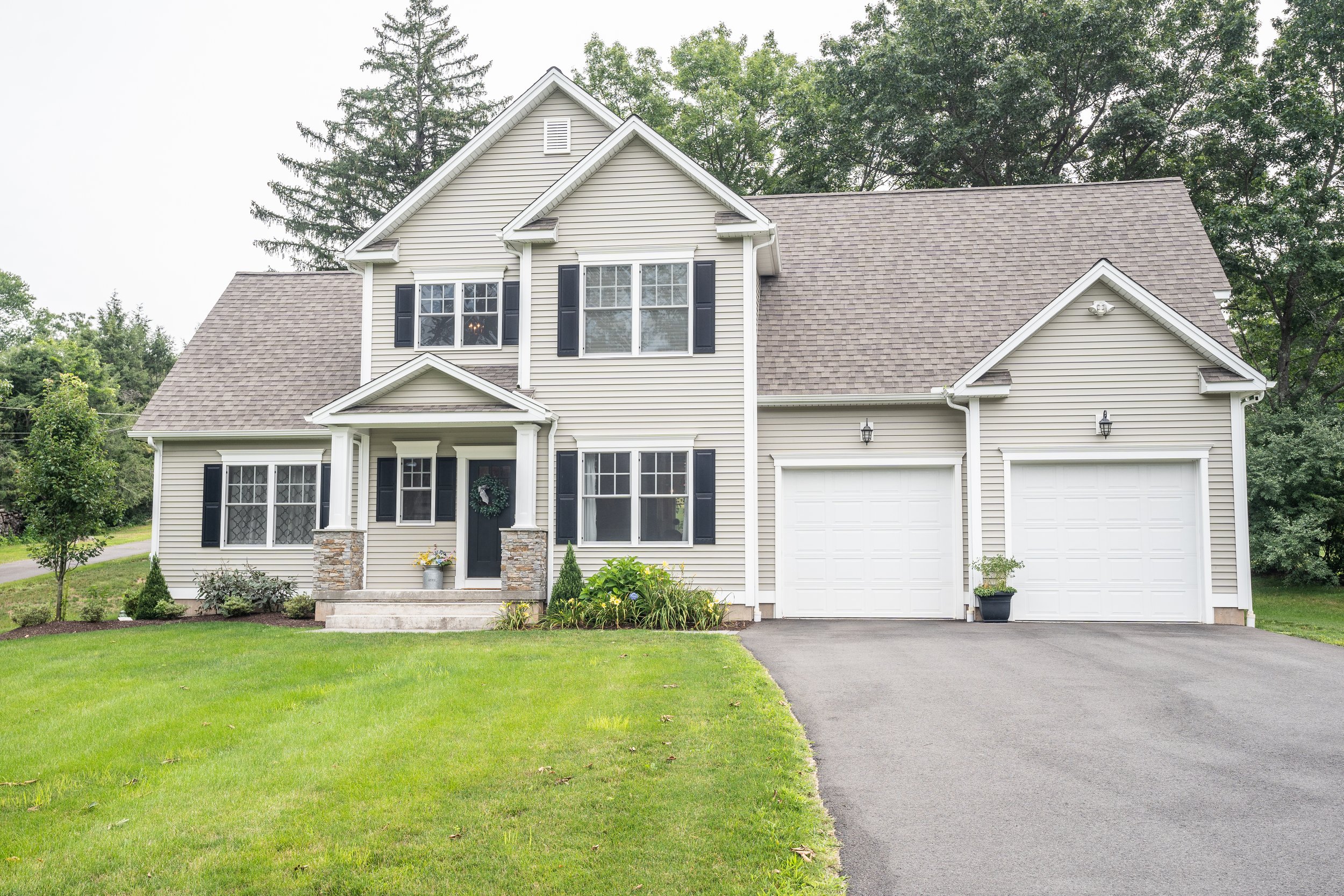 A two-story suburban house with beige siding, black shutters, a front porch with stone columns, and a garage with two white doors, surrounded by a green lawn and trees in the background.