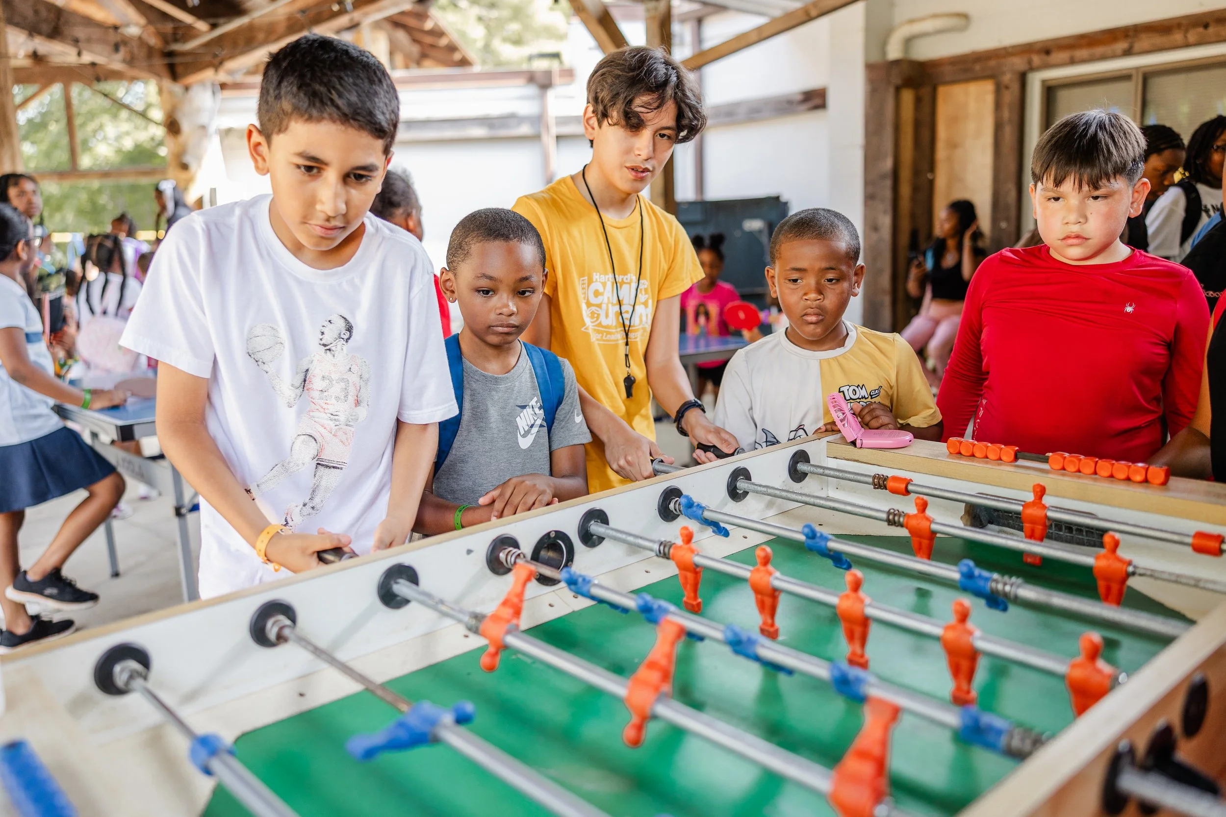 Six children playing foosball together in an indoor recreation space with a wooden roof and several people in the background.