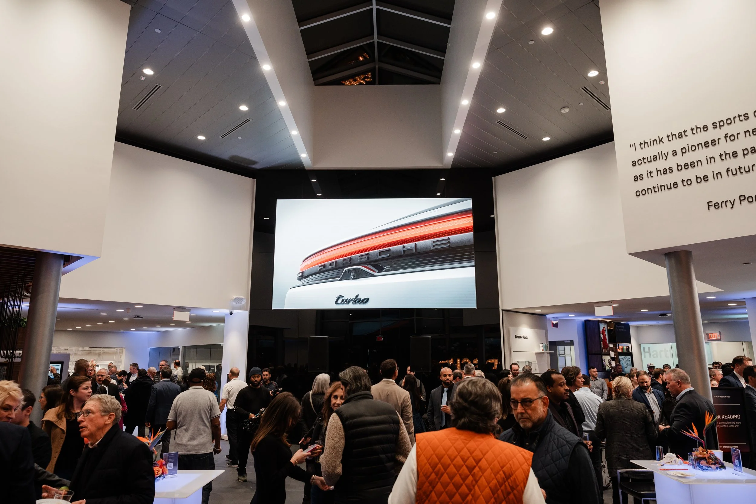 A crowded event space with many people mingling and socializing. A large digital screen displaying a Porsche car with the words 'turbo' is at the center. The room has modern architectural design with white walls, a high ceiling with recessed lighting