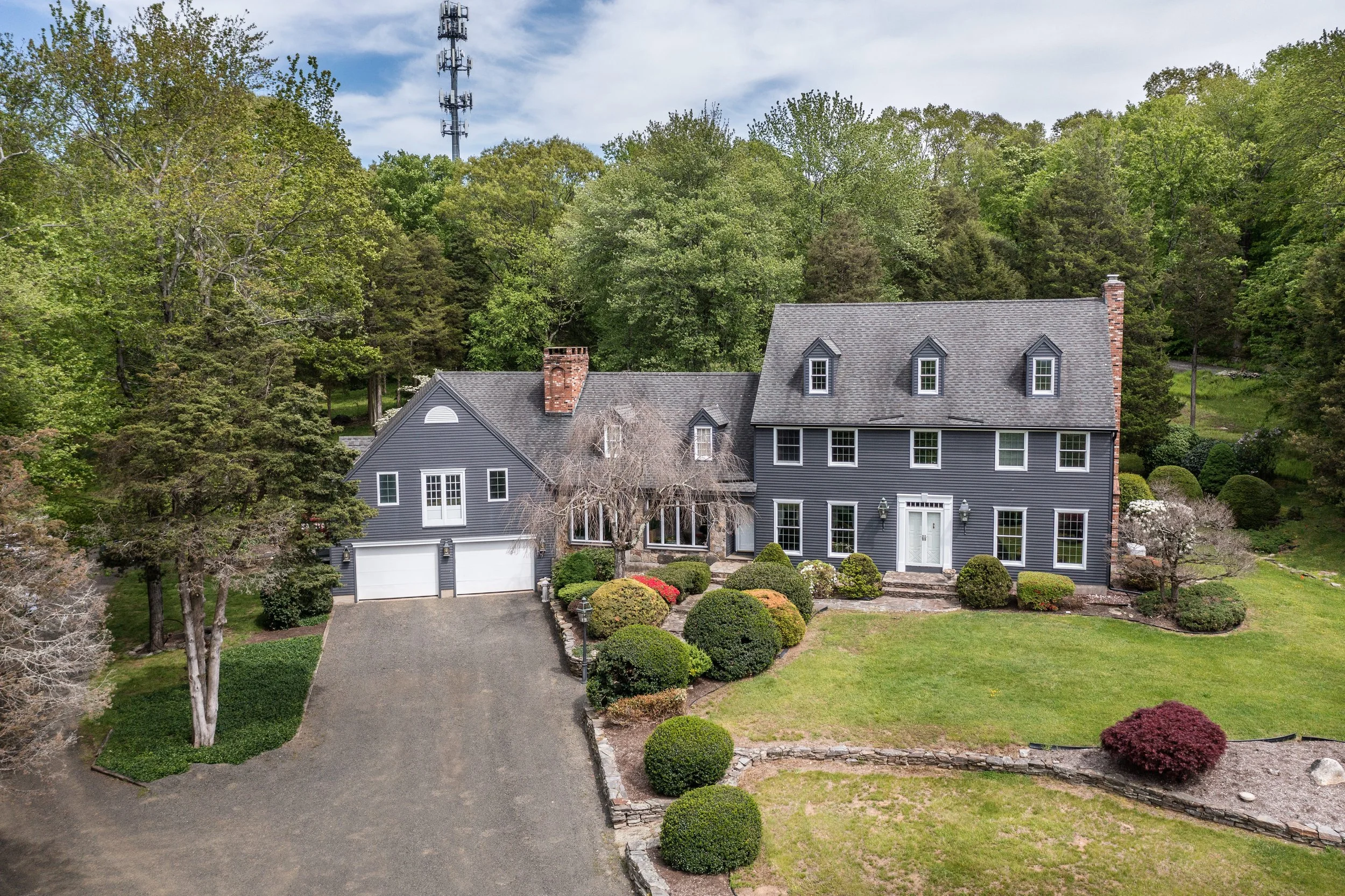 A large gray house with multiple windows and a front door, surrounded by a well-maintained lawn and bushes, with a driveway leading to a garage, set against a backdrop of trees under a partly cloudy sky.